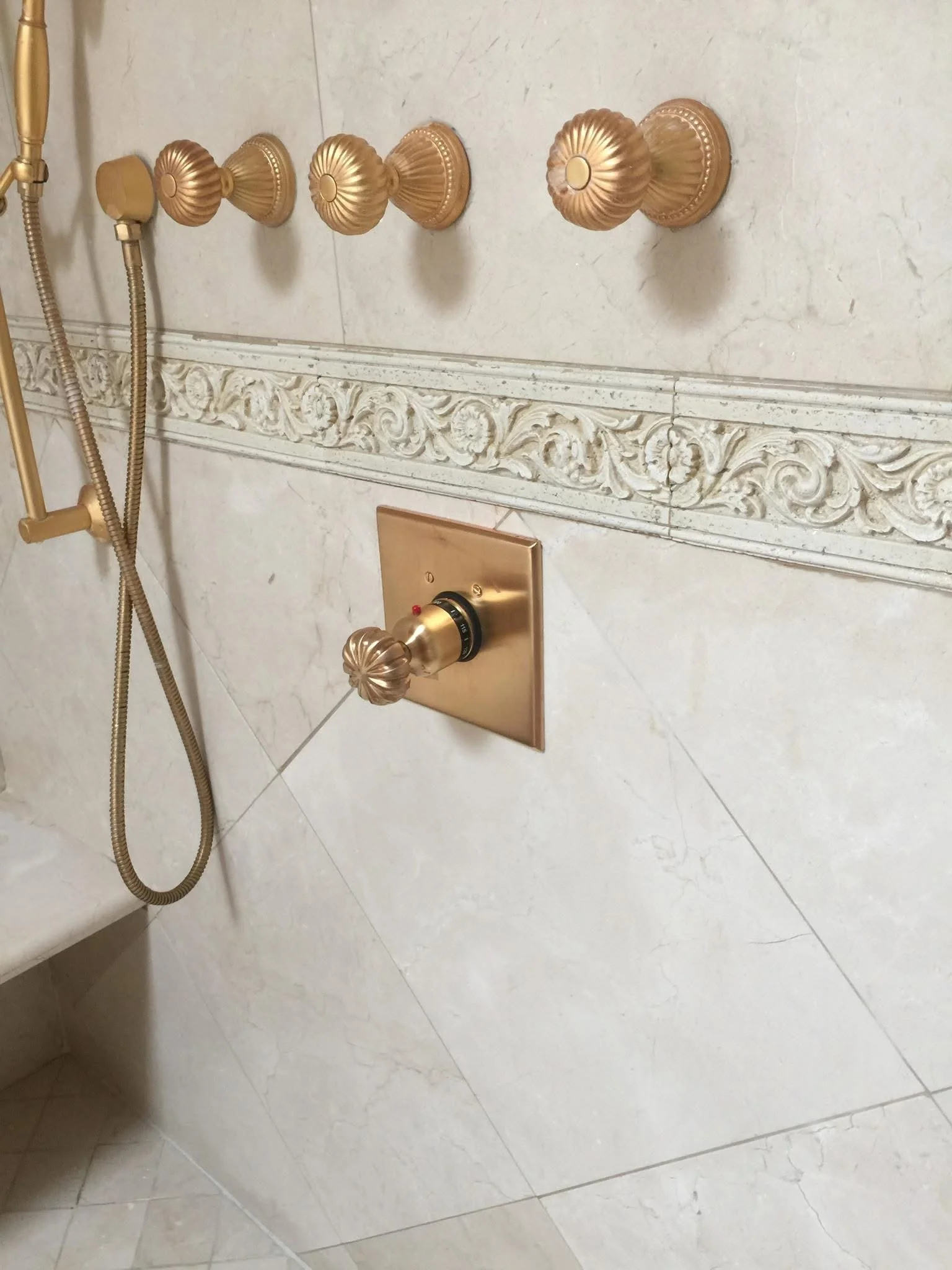 Close-up of a luxurious shower wall with gold-colored fixtures, including multiple knobs, a showerhead, and a temperature control dial, set against a marble wall with decorative molding.