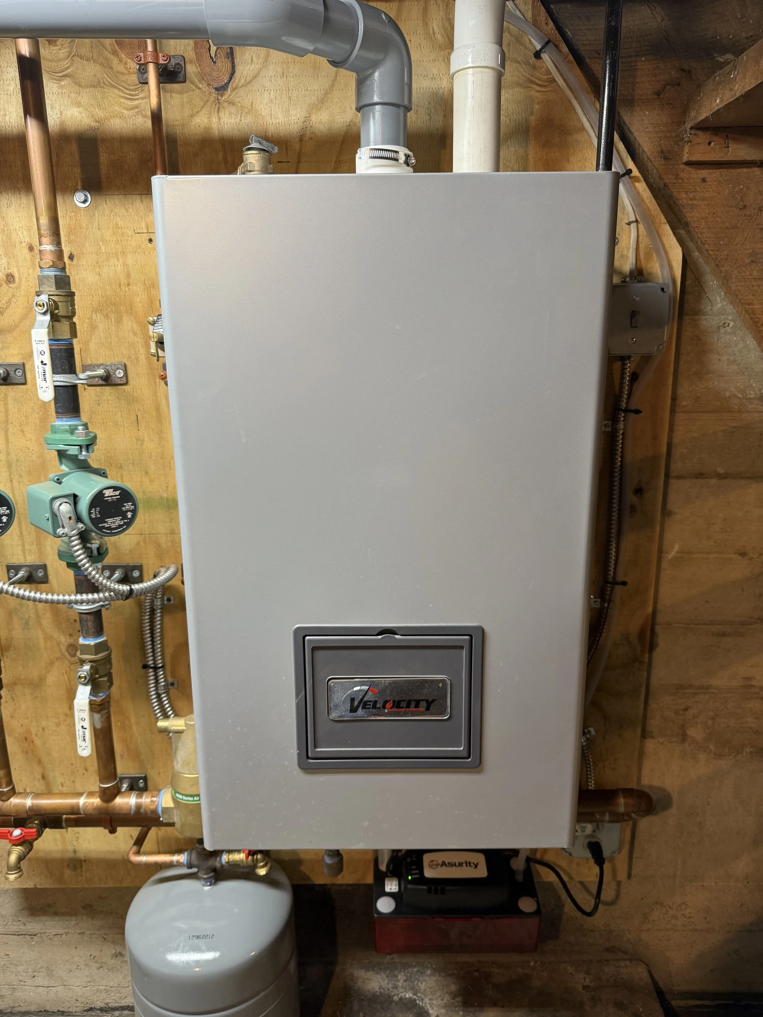 Image of a gray Velocity tankless water heater mounted on a wooden wall, with copper and plastic pipes connected on the left and a small device labeled Asurity below it.