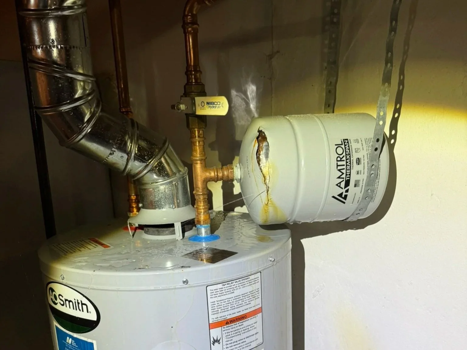 A water heater with a large crack and water leaking from it, connected to pipes and a yellow expansion tank, in a utility room.