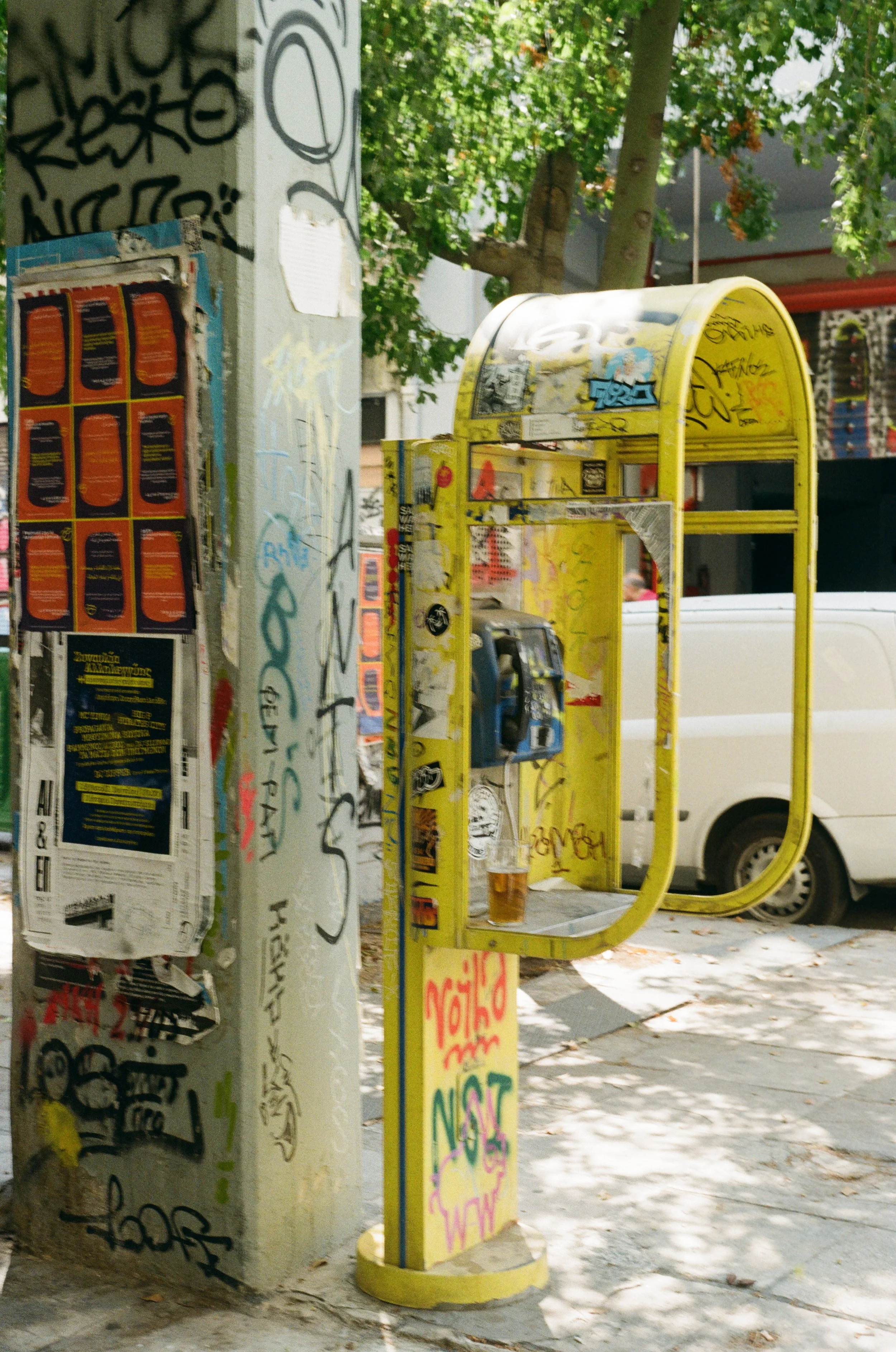 A yellow public payphone booth covered in graffiti on a sidewalk, with a white van parked and trees with green leaves in the background.