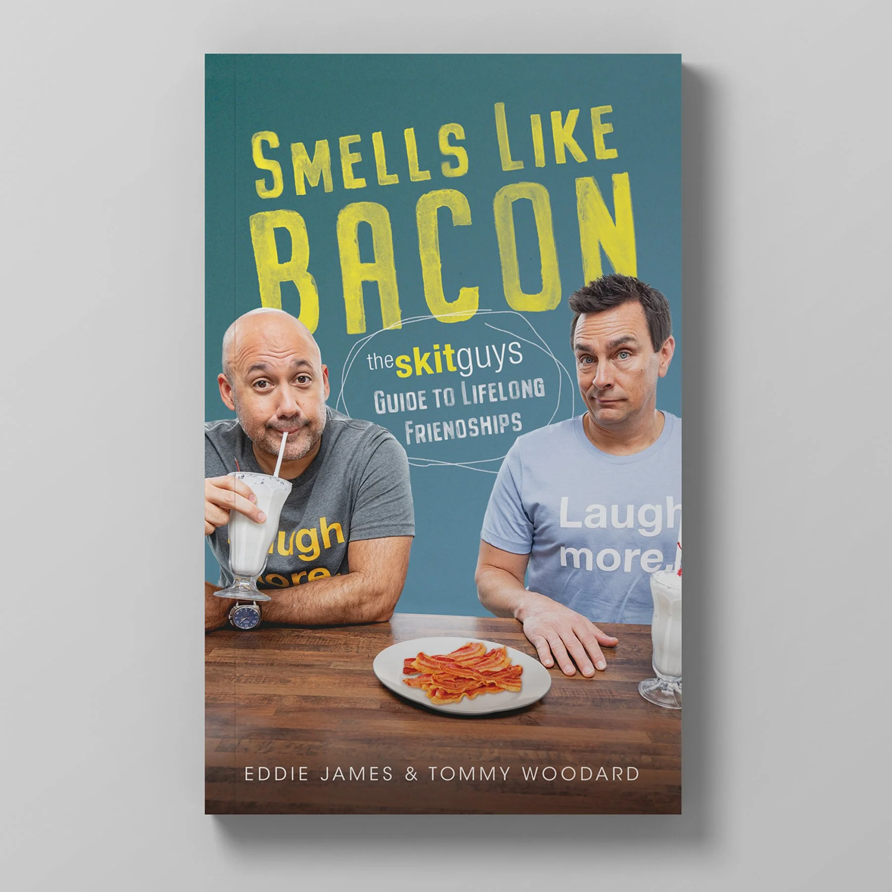Book cover titled 'Smells Like Bacon' featuring two men, one with a milkshake, and the other pointing at bacon on a plate, with a blue background.