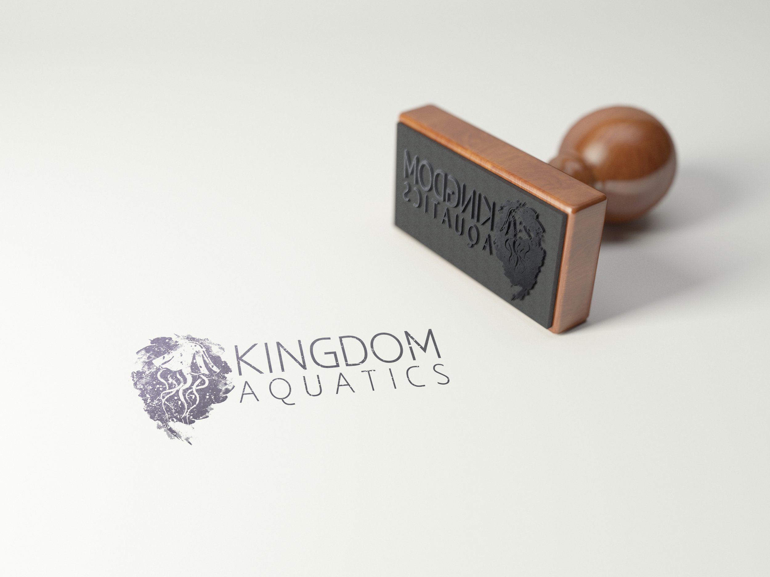 A rubber stamp marked with the Kingdom Aquatics logo, resting on a white surface next to a printed logo of the same design.