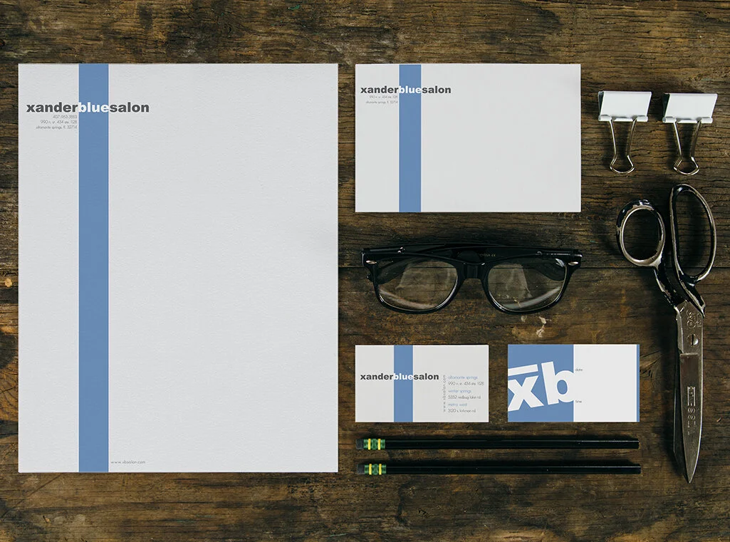 Business stationery and office supplies from Xander Blue Salon, including letterhead, appointment card, business card, envelopes, black glasses, scissors, two binder clips, two pencils, on a wooden surface.