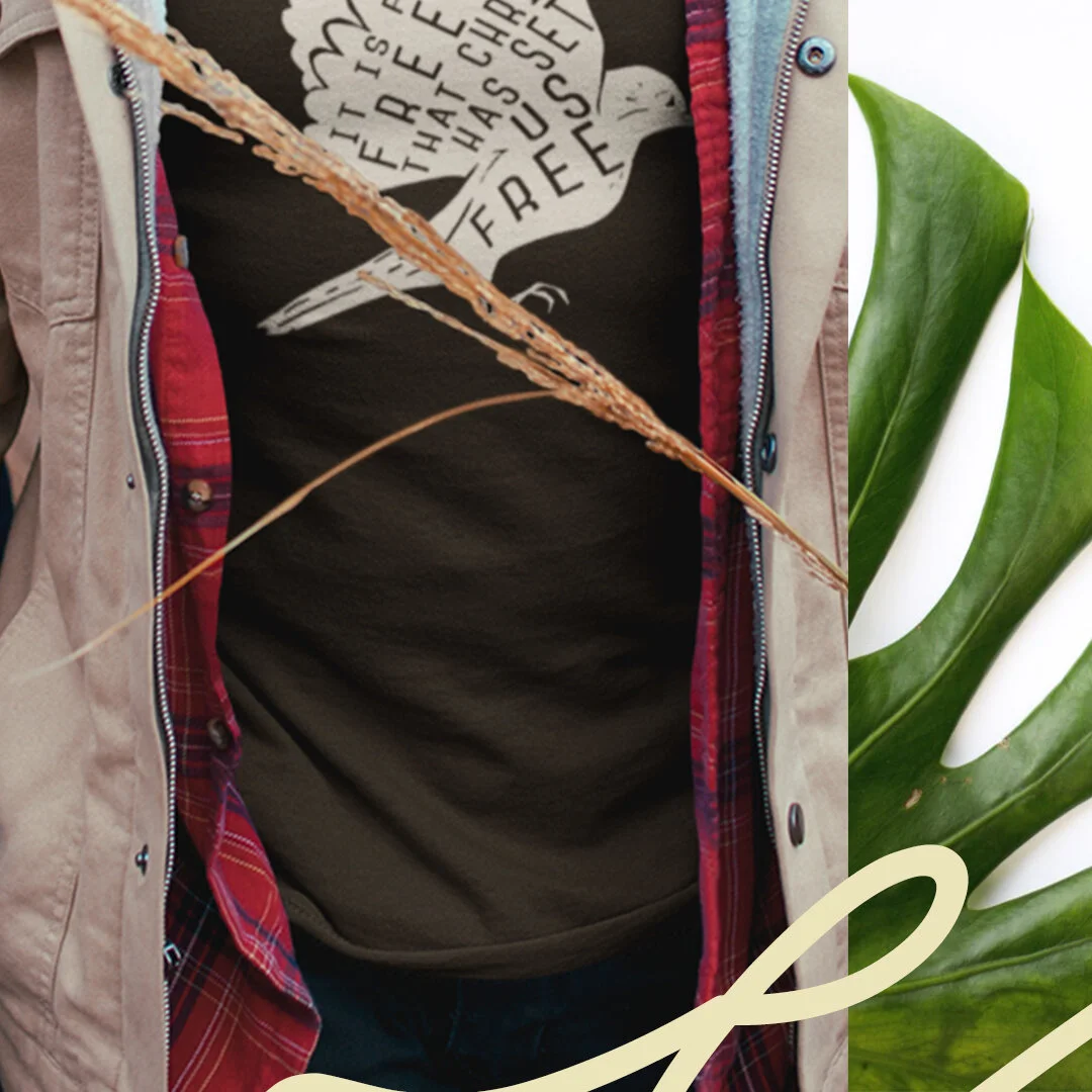 Part of a person wearing a black t-shirt with a white bird graphic and text, a red plaid shirt, and a tan jacket, with dried high grass in the foreground and large green leaves on the side.