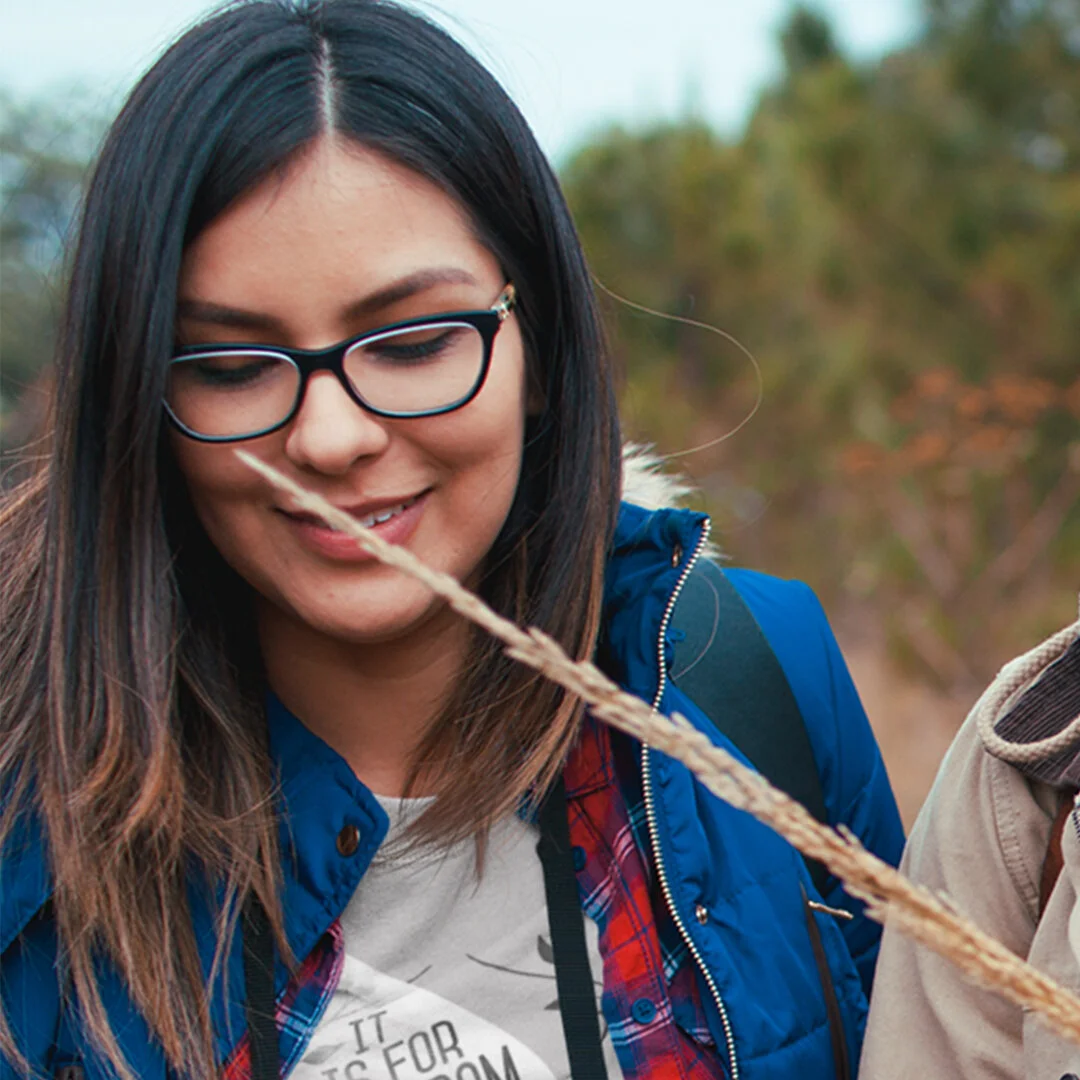 Young woman with long dark hair and glasses smiling outdoors, wearing a blue jacket and a plaid shirt.