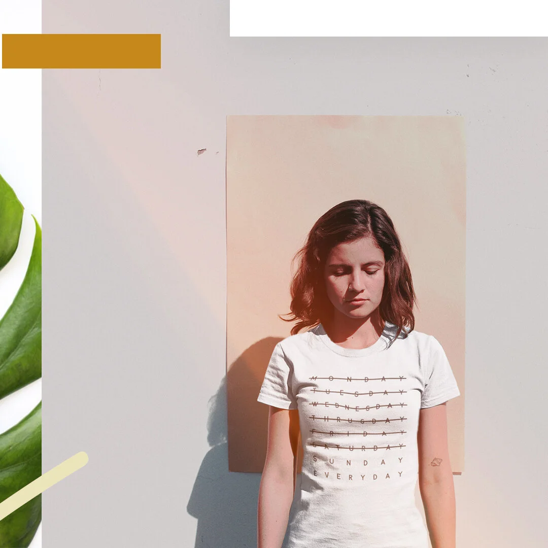 A young woman with shoulder-length brown hair standing against a beige background with sunlight casting shadows, wearing a white graphic t-shirt with days of the week written on it.
