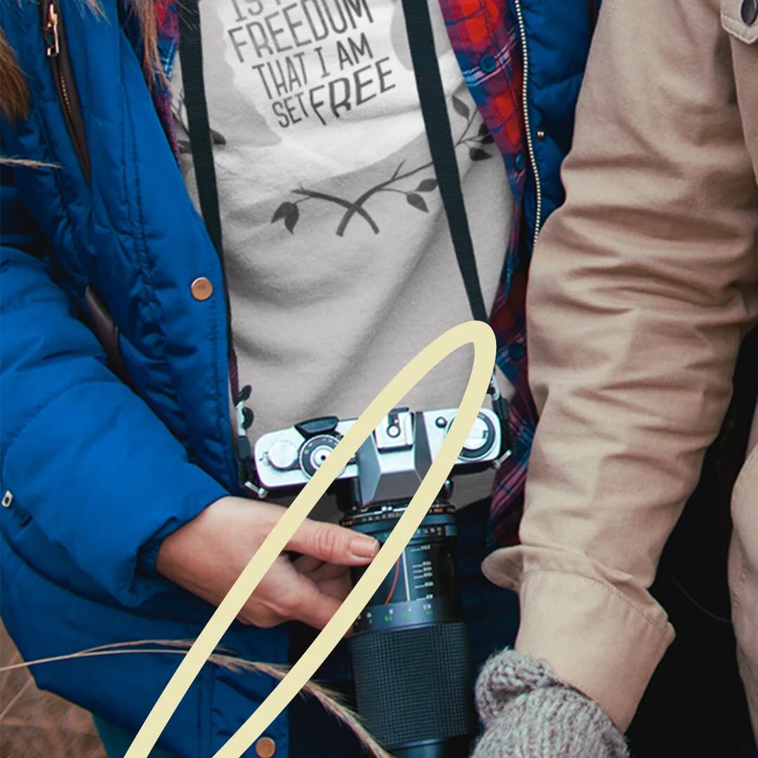 Close-up of two people, one holding a camera on a strap, person on the left wearing a blue jacket and beige shirt with a quote, person on the right in a beige jacket and gray knitted glove.