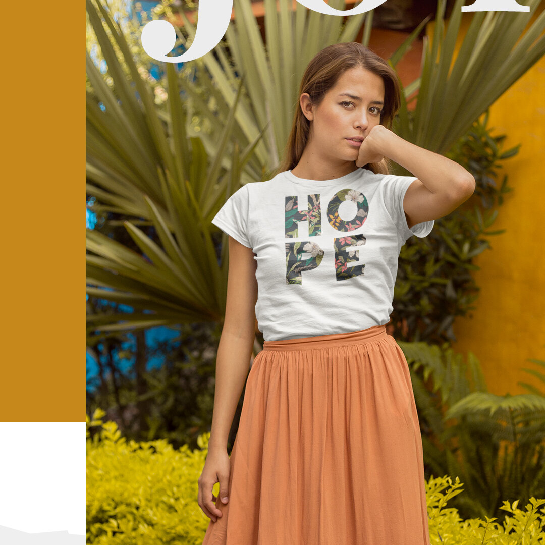 A young woman with brown hair wearing a white t-shirt with floral letters spelling "HOPE" and a long orange skirt, standing outdoors in front of large green plants.