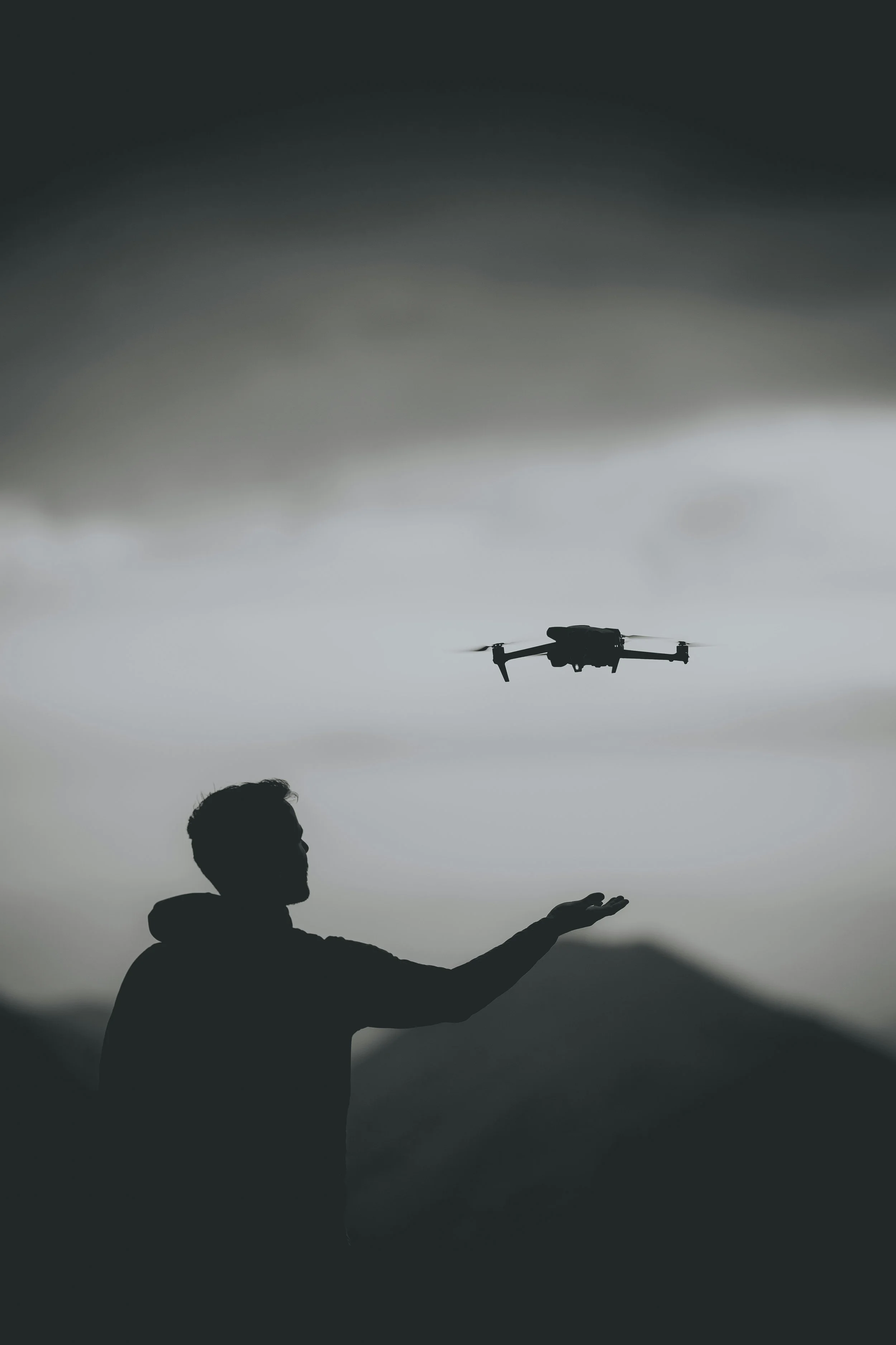Silhouette of a person with outstretched arm and a drone flying above against a cloudy sky.