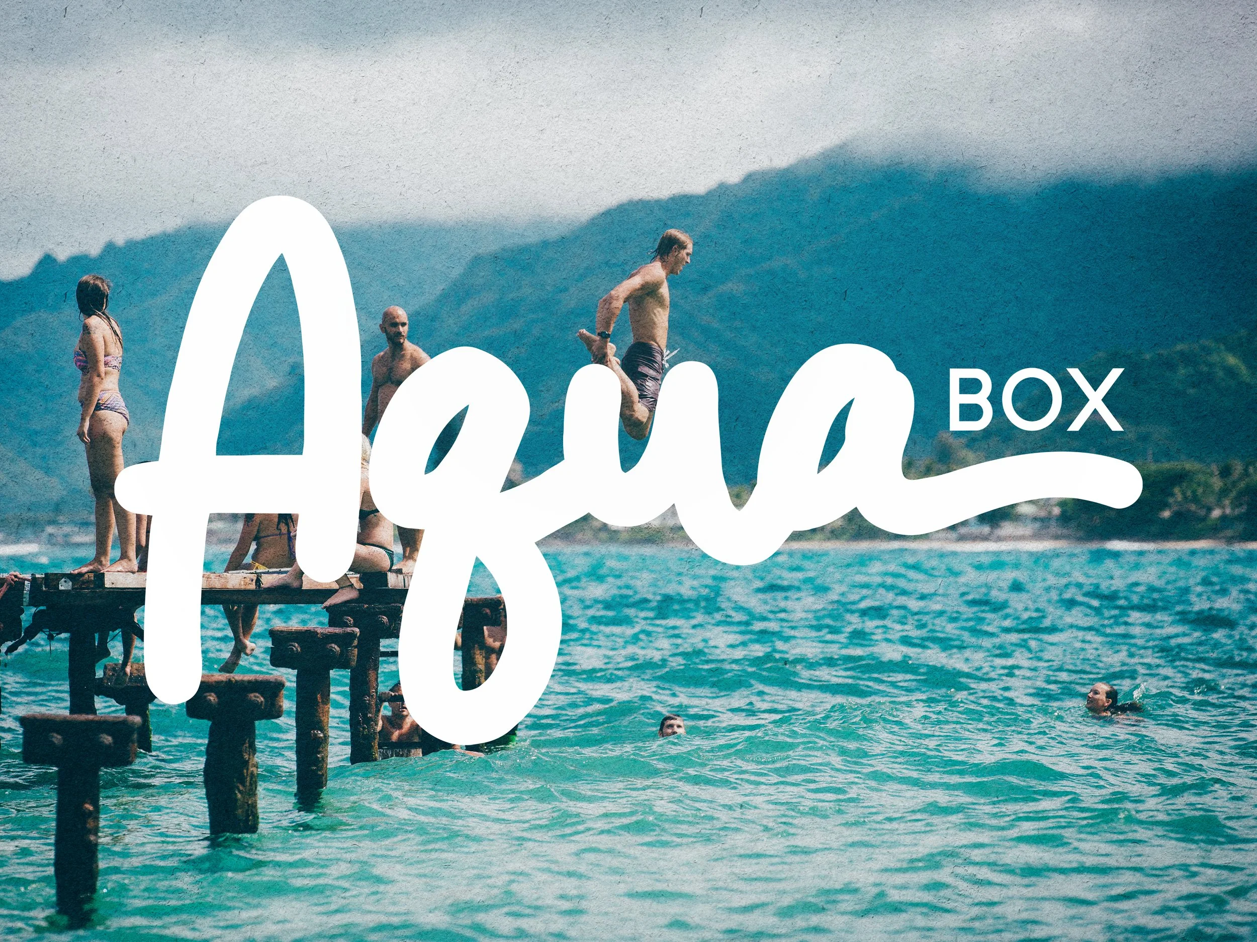 People jumping into the ocean from a wooden pier on a sunny day with mountains in the background