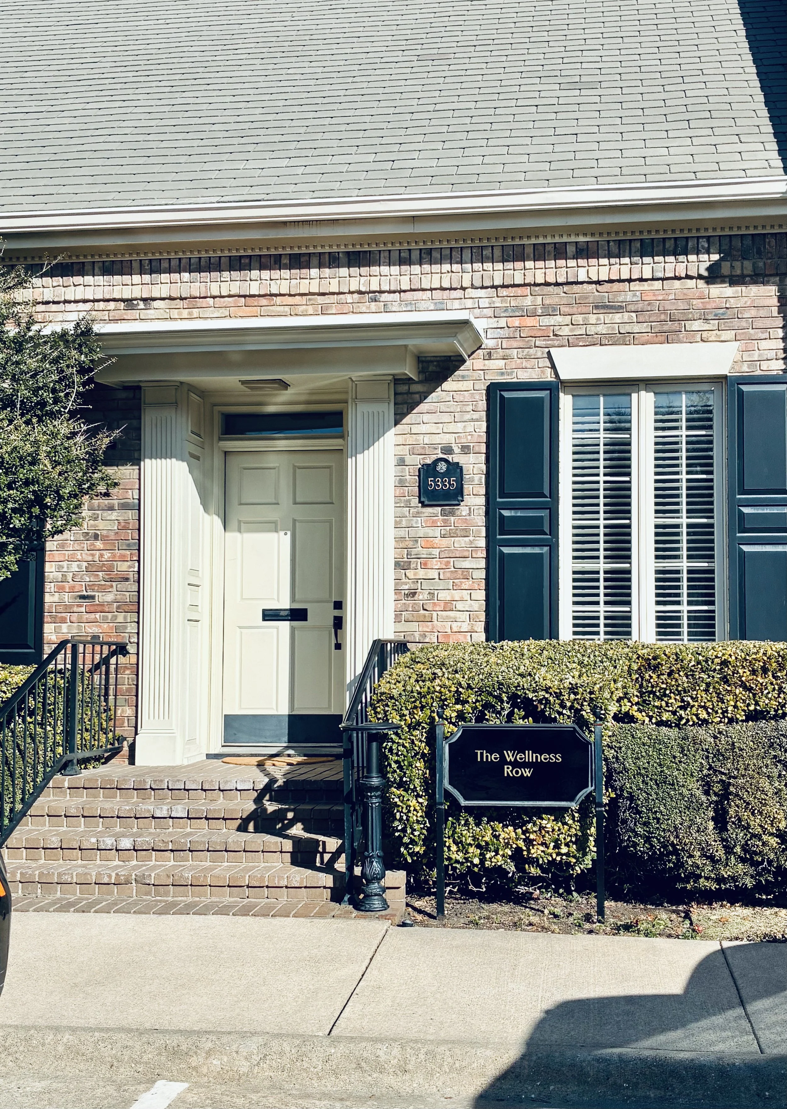 A wellness collective that is part of wellness row with brick exterior, white door, black and white trim, and a sign reading "The Wellness Row" in front of neatly trimmed bushes.