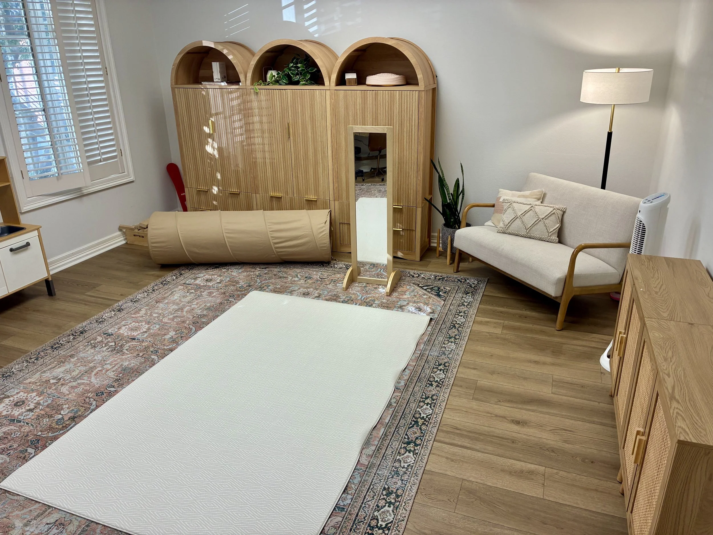 Room with large window, wooden furniture including a wardrobe, a small sofa with pillows, a standing floor lamp, a mirror, and a patterned rug on hardwood flooring.