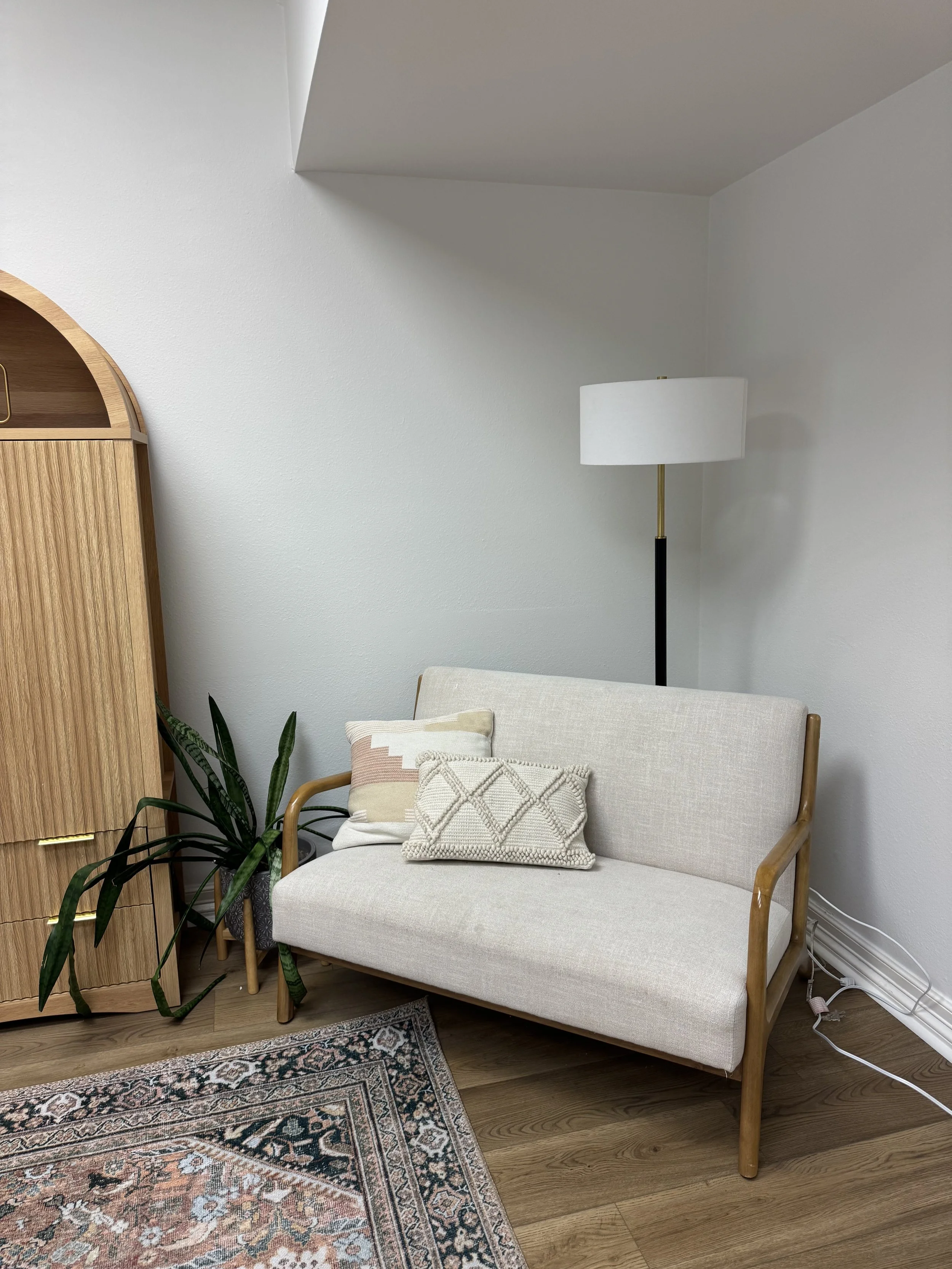 A beige sofa with two decorative pillows, a potted plant, a wooden cabinet, a patterned rug, and a floor lamp in a cozy physical therapy room that parents are able to sit on while in child is improving on gross motor skills.