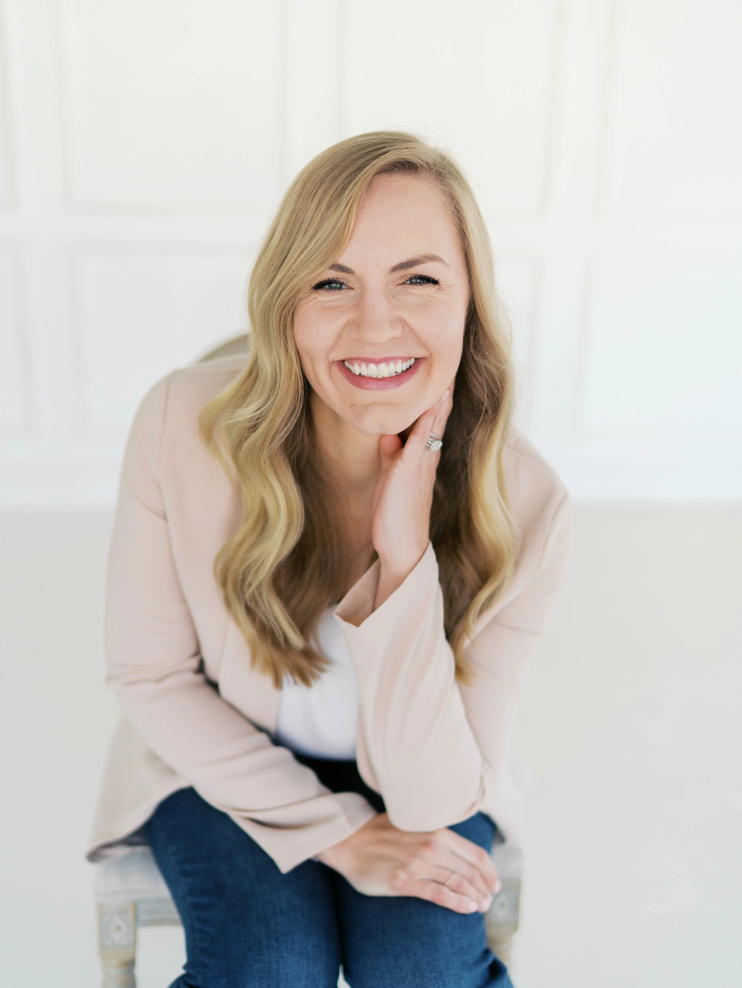 A smiling female, holistic, wellness based physical therapist with blonde hair, wearing a light blazer and dark jeans, sitting on a chair in a minimalist physical therapy treatment room.
