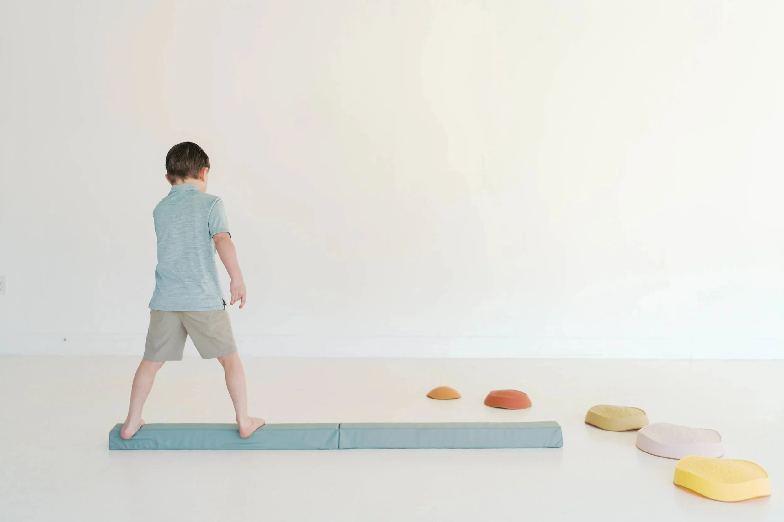 A young boy participating in physical therapy for balance and coordination impairments. He is also participating in foot strengthening activities, with colorful stepping stones placed on the floor nearby in a comforting non-toxic environment.