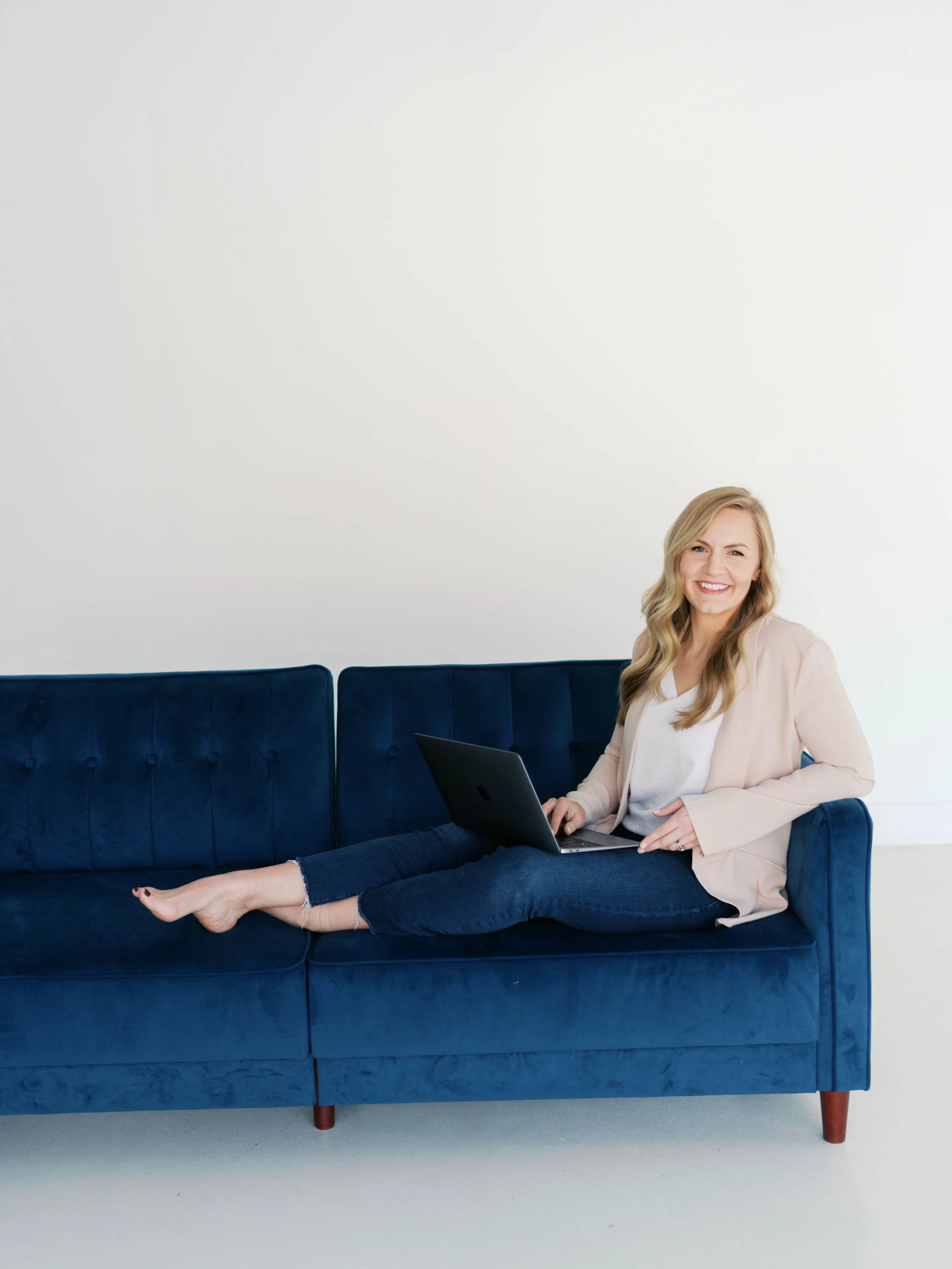 A woman with blonde hair wearing a beige blazer and white shirt, sitting on a dark blue sofa, holding a laptop, smiling in a bright room with a white wall.