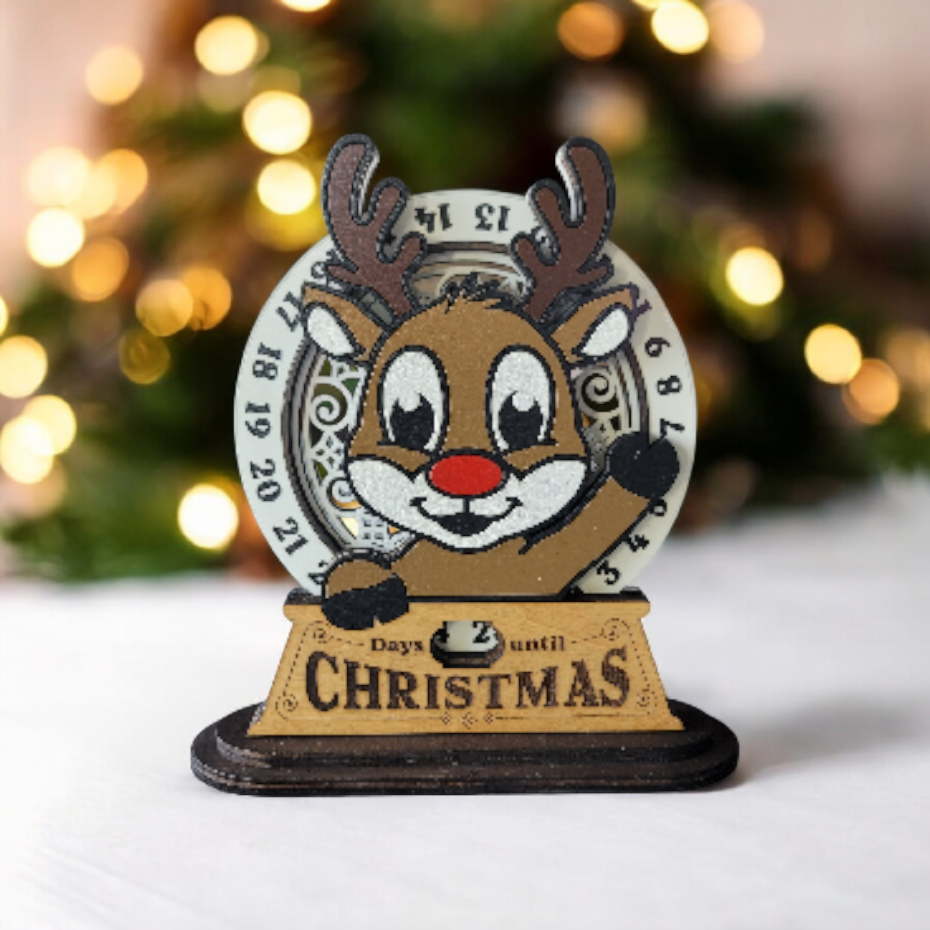 Decorative reindeer figure with a red nose and black gloves, surrounded by a circular countdown and a sign that reads "2 days until Christmas," against a blurred Christmas tree with lights in the background.