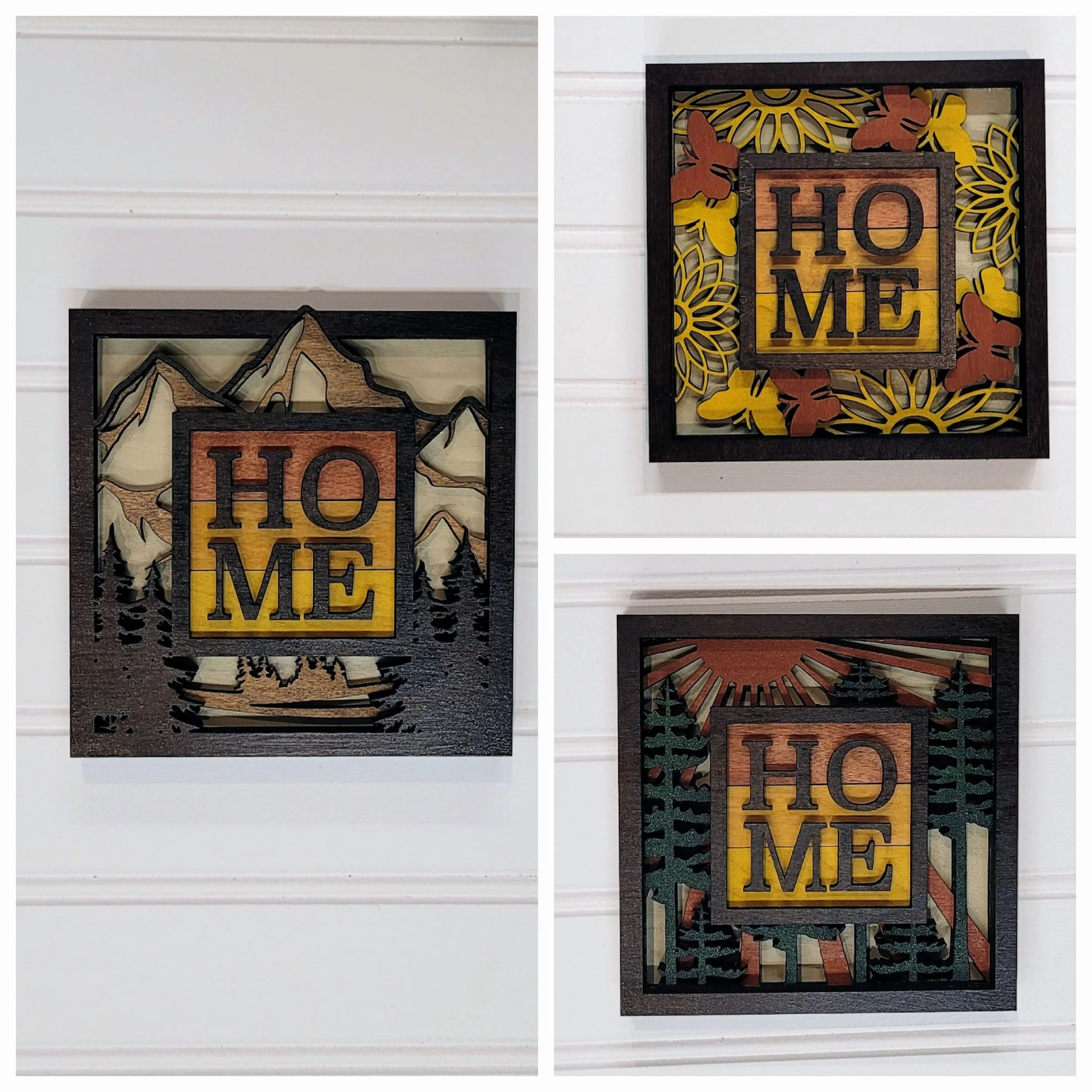 Three wall art pieces with the word 'HOME' in the center. The left piece features a mountain landscape with pine trees. The top right piece has a sunflower and butterfly design. The bottom right piece shows a forest scene with the rising or setting s