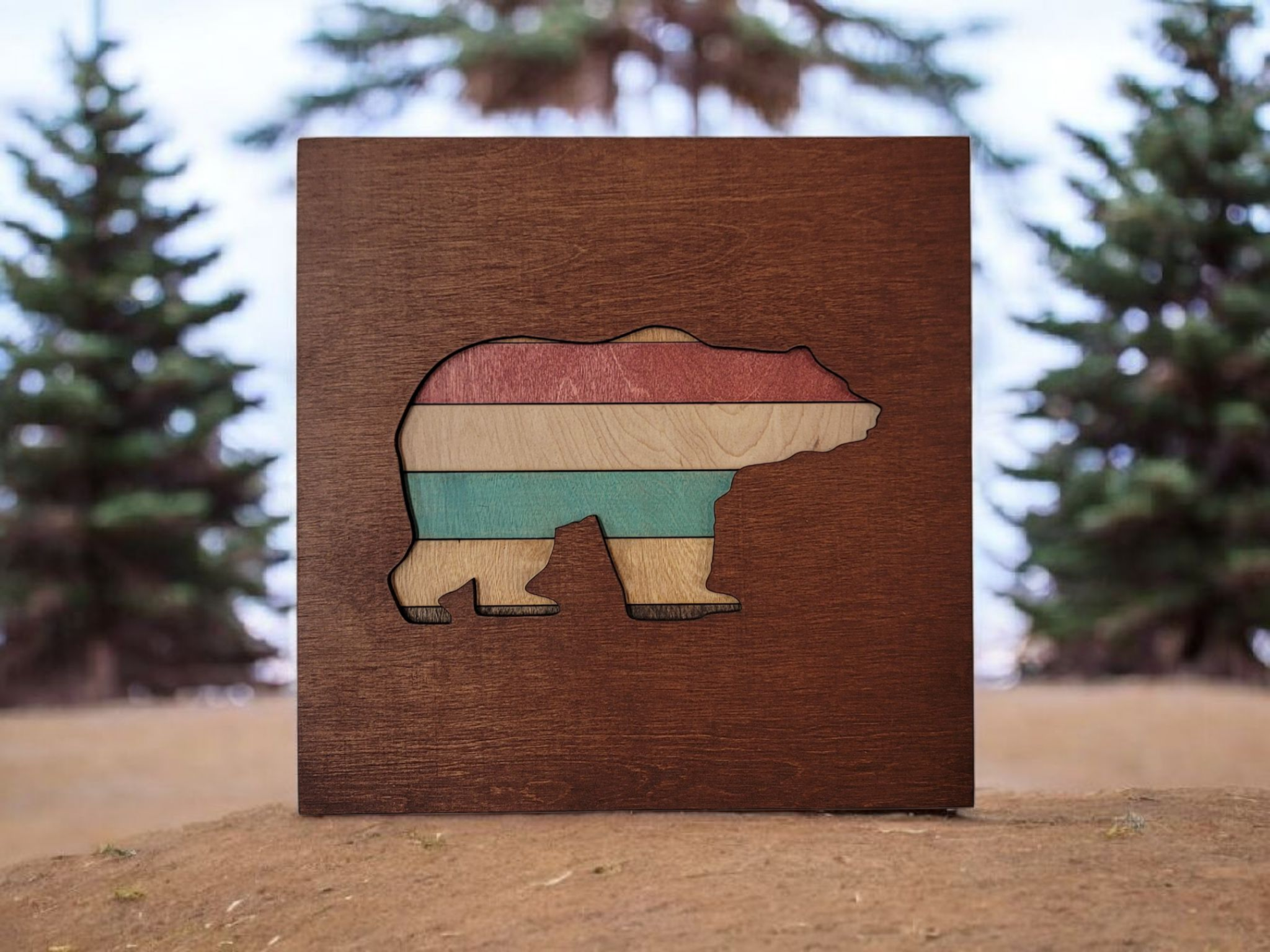 Wooden art piece in the shape of a bear with horizontal stripes of red, beige, and teal on a wooden background, outdoors with evergreen trees in the blurred background.