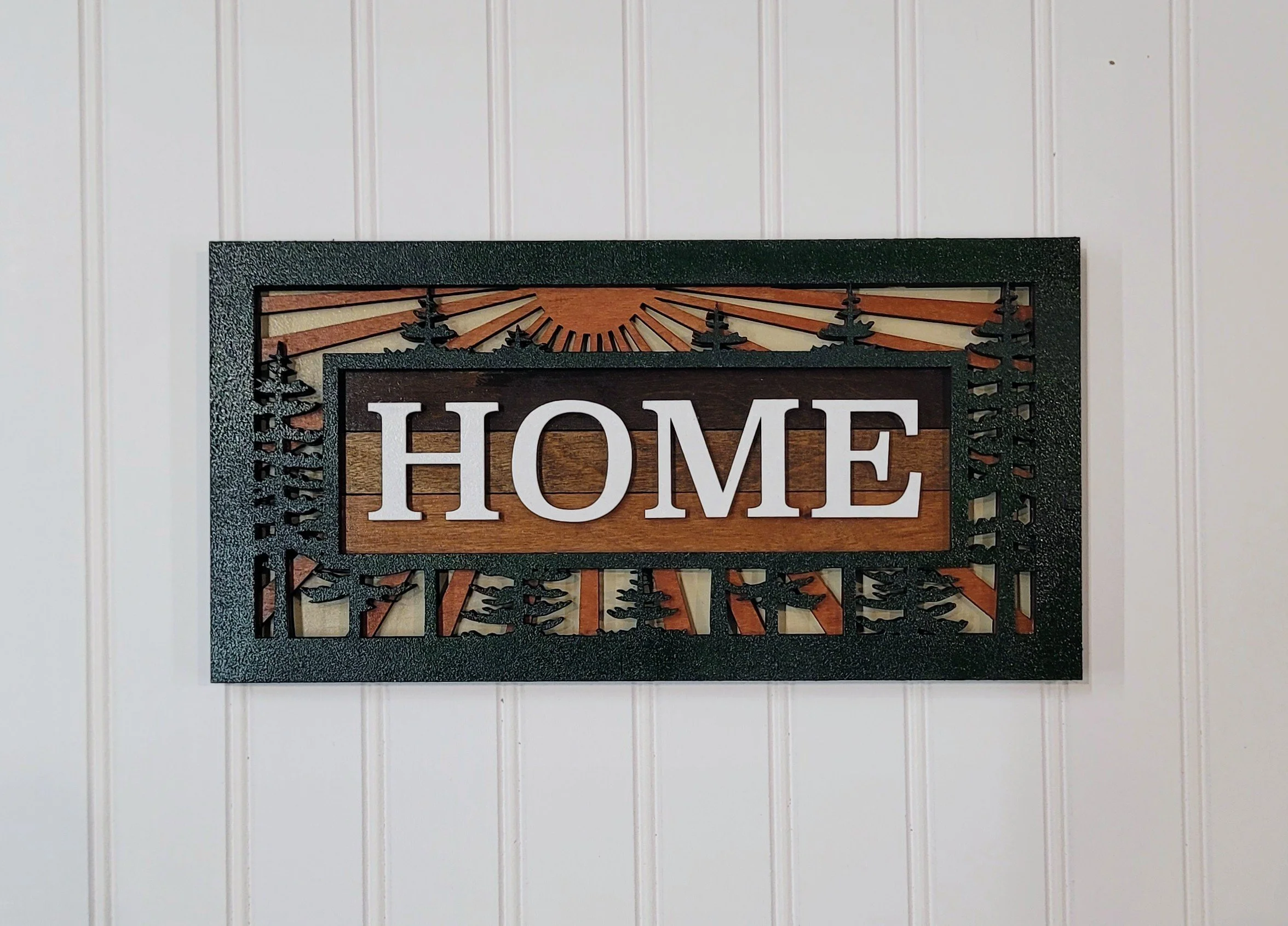 Wall-mounted decorative sign with the word 'HOME' in white letters and a nature scene background featuring trees and a sunrise or sunset.