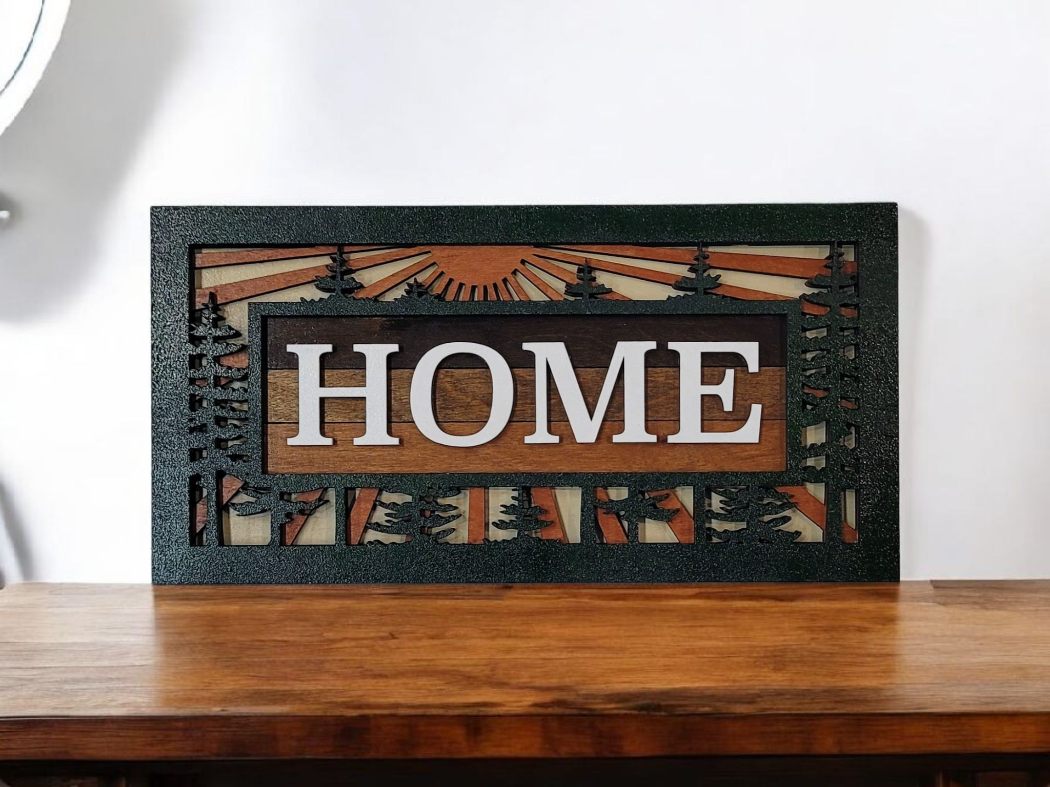 Decorative wooden wall art with the word "HOME" in large white letters, surrounded by a frame with tree silhouettes and a sunrise pattern.