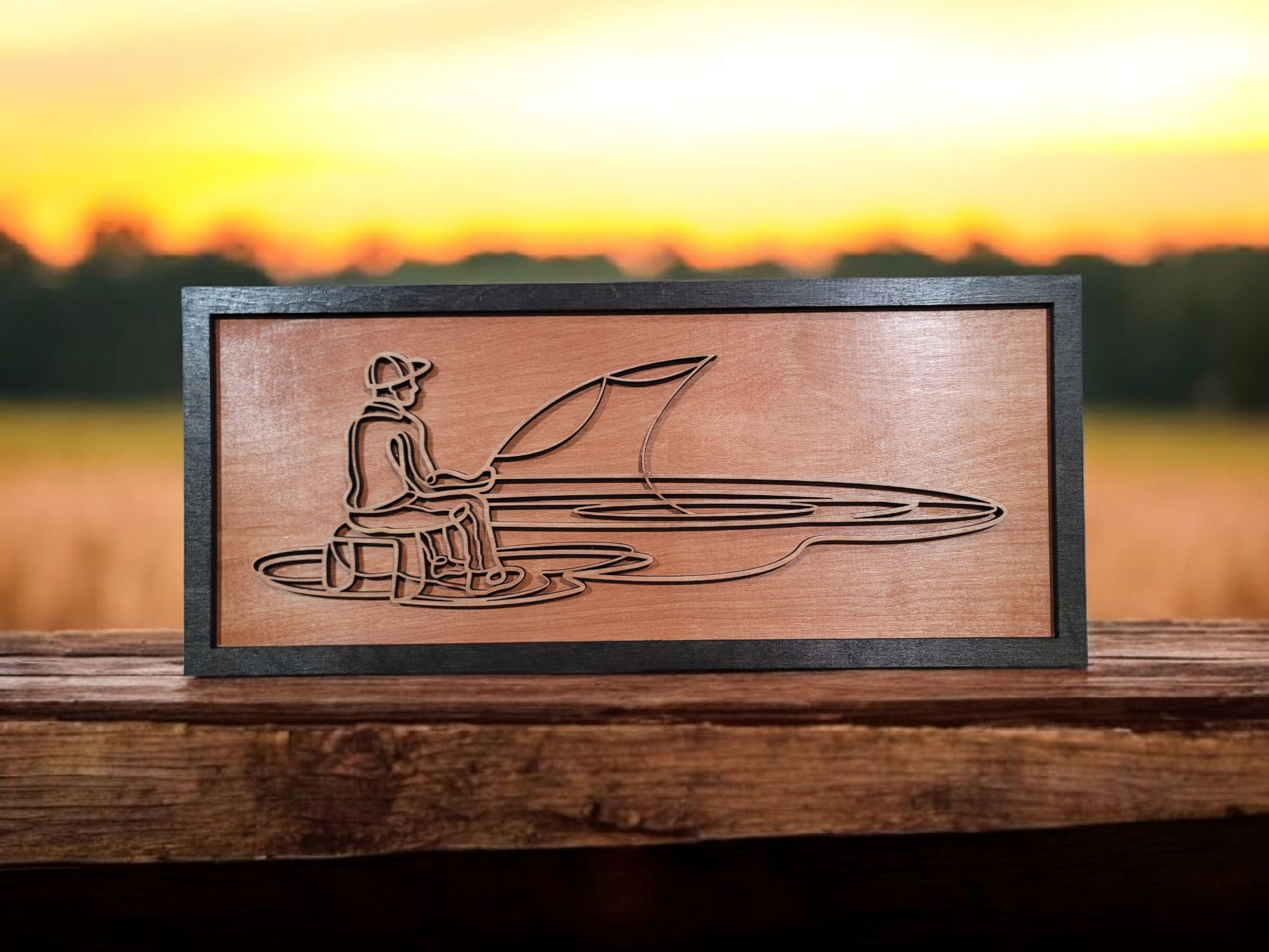 Wooden artwork of a person sitting on a bench with a cap, holding a fishing rod next to a sailboat, placed on a wooden surface outdoors at sunset.
