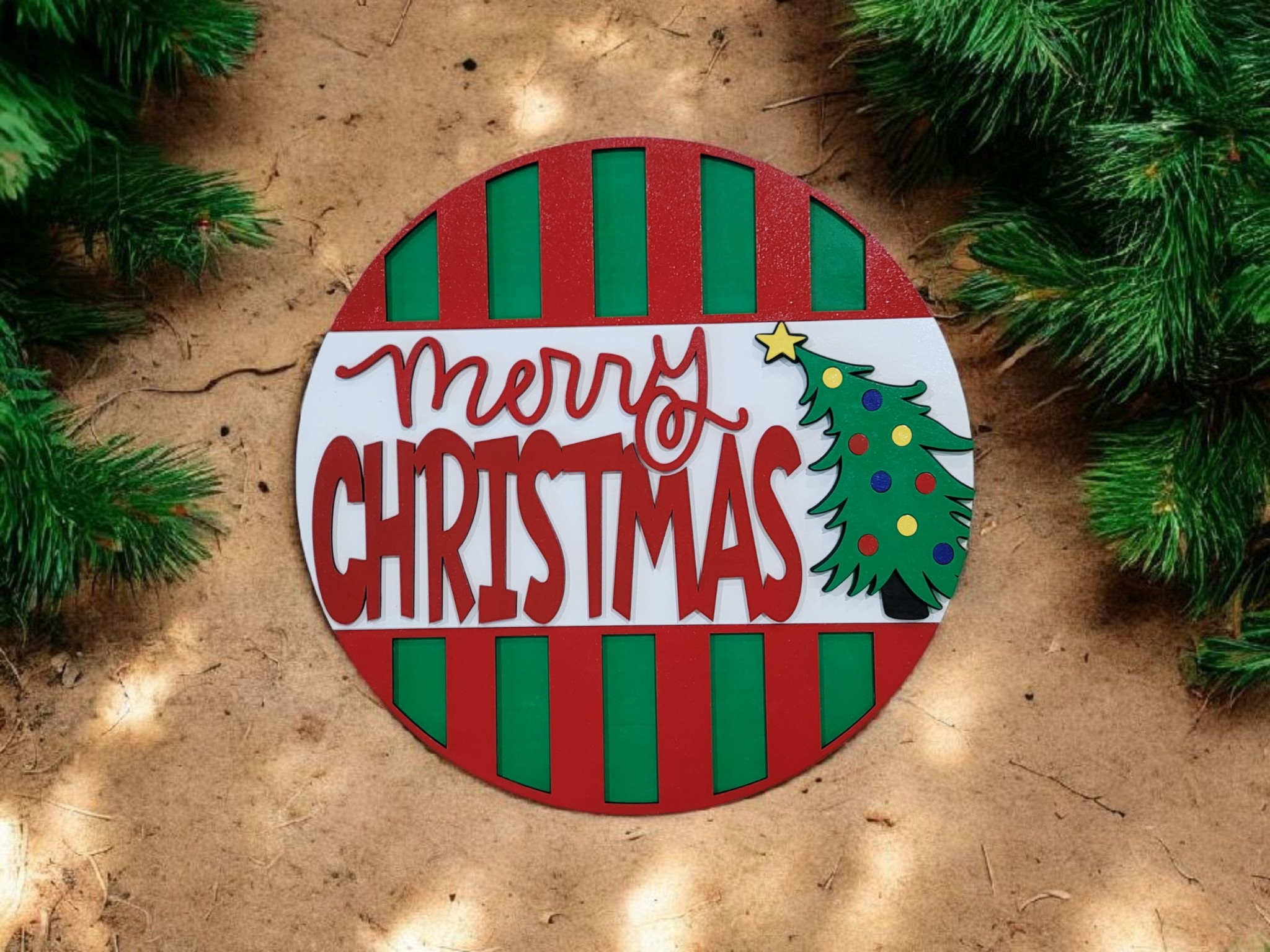 Decorative Christmas sign with the message 'Merry Christmas' next to a Christmas tree illustration, surrounded by green pine branches on a sandy background.