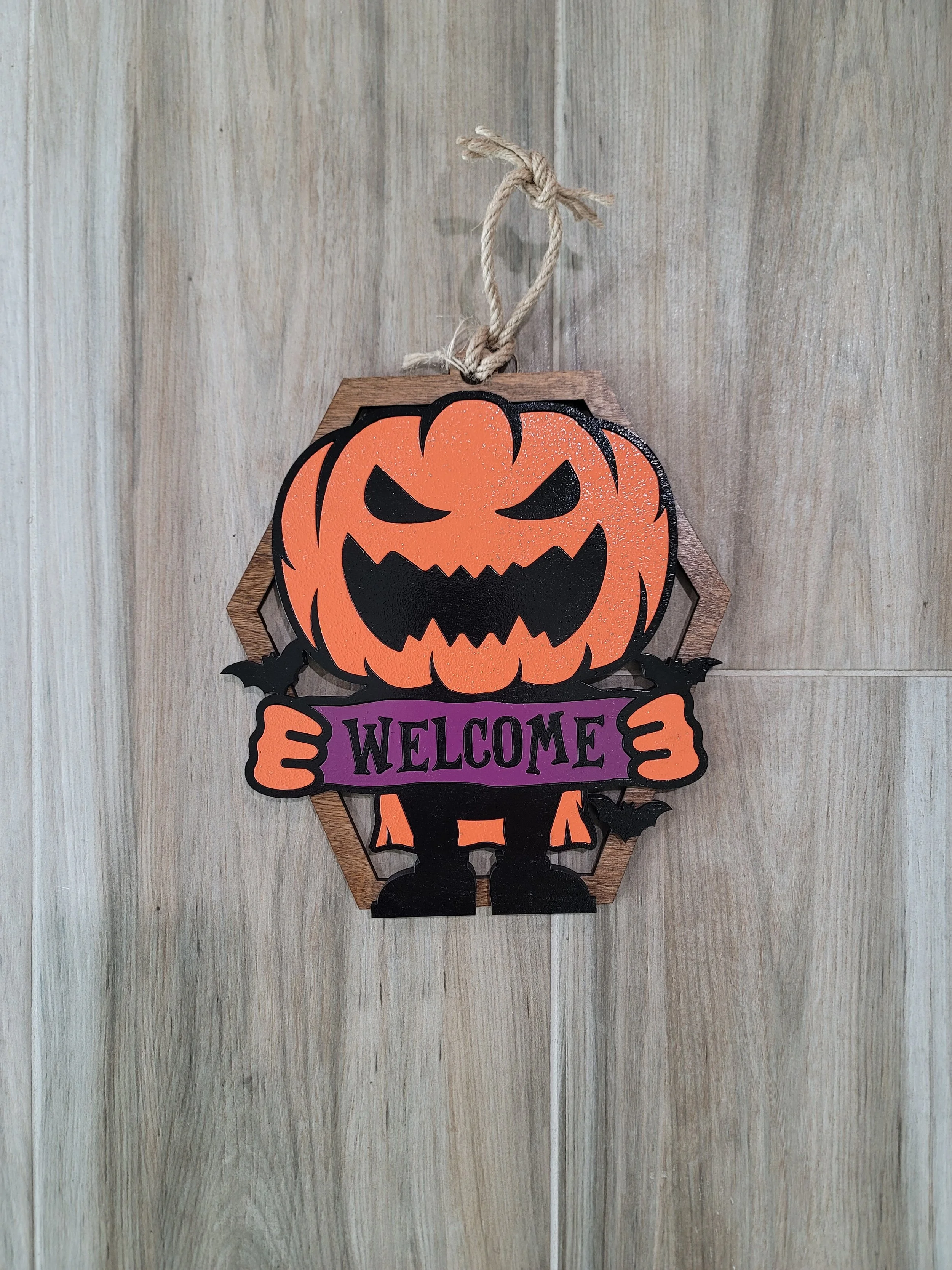 A Halloween-themed decorative sign featuring a cartoon pumpkin with a menacing face, holding a purple sign that says 'WELCOME'. The pumpkin and sign are mounted on a hexagonal wooden background with a jute string for hanging.