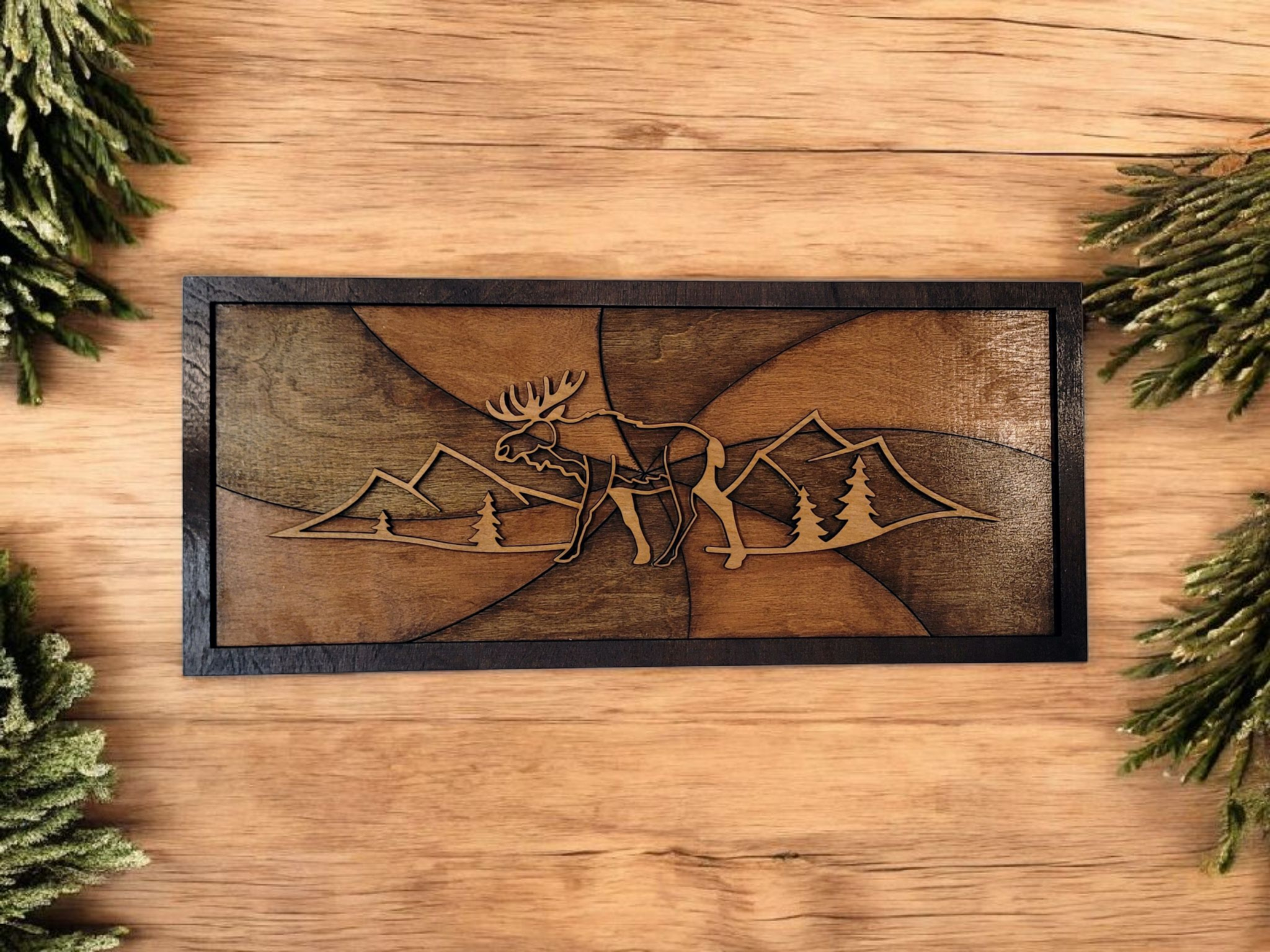 Wooden wall art with a mountain and pine trees scene, featuring a large moose in the center, framed in black, placed on a wooden surface with pine branches around it.