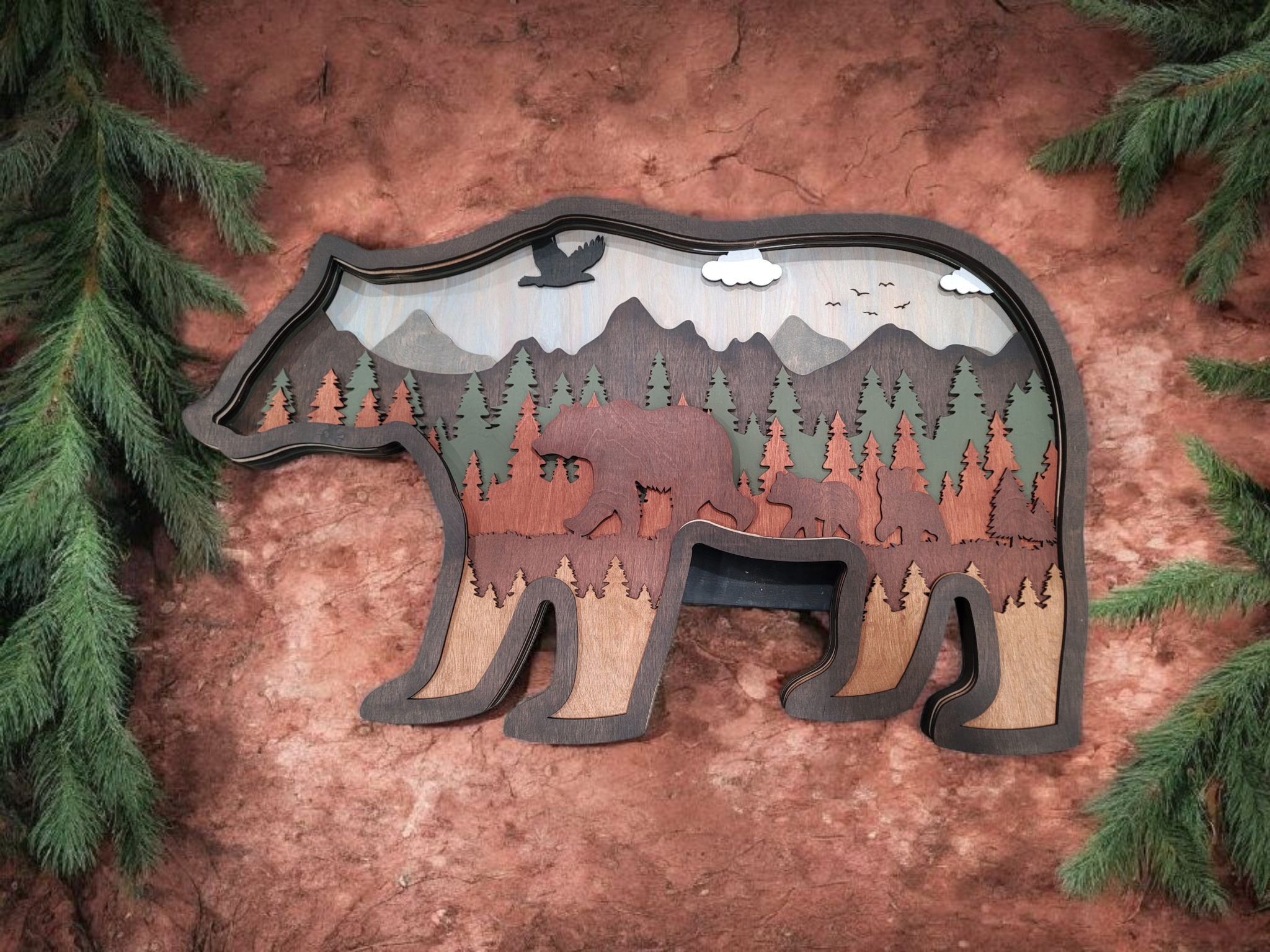 Wooden bear-shaped wall art with forest, mountains, and wildlife scene, surrounded by green pine branches on red dirt ground.
