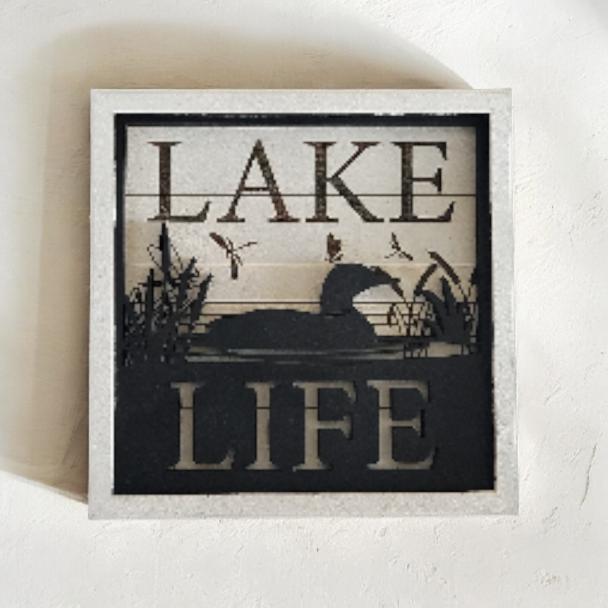 Decorative wall art with the words 'LAKE LIFE' and a silhouette of a duck on a lake with reeds and flying birds.