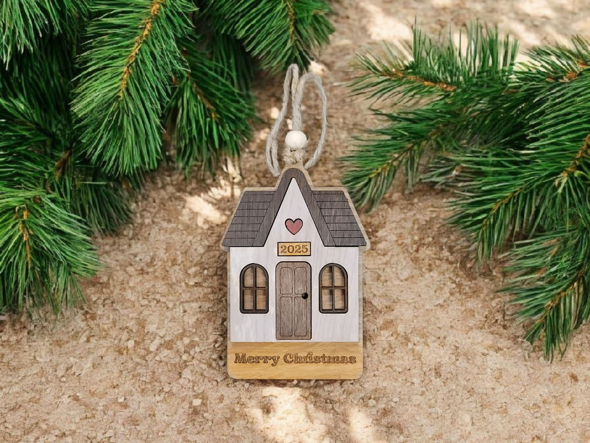 Wooden Christmas ornament in the shape of a house with a heart, the year 2025, and the words "Merry Christmas" engraved, hanging from a string amidst pine branches on a sandy surface.