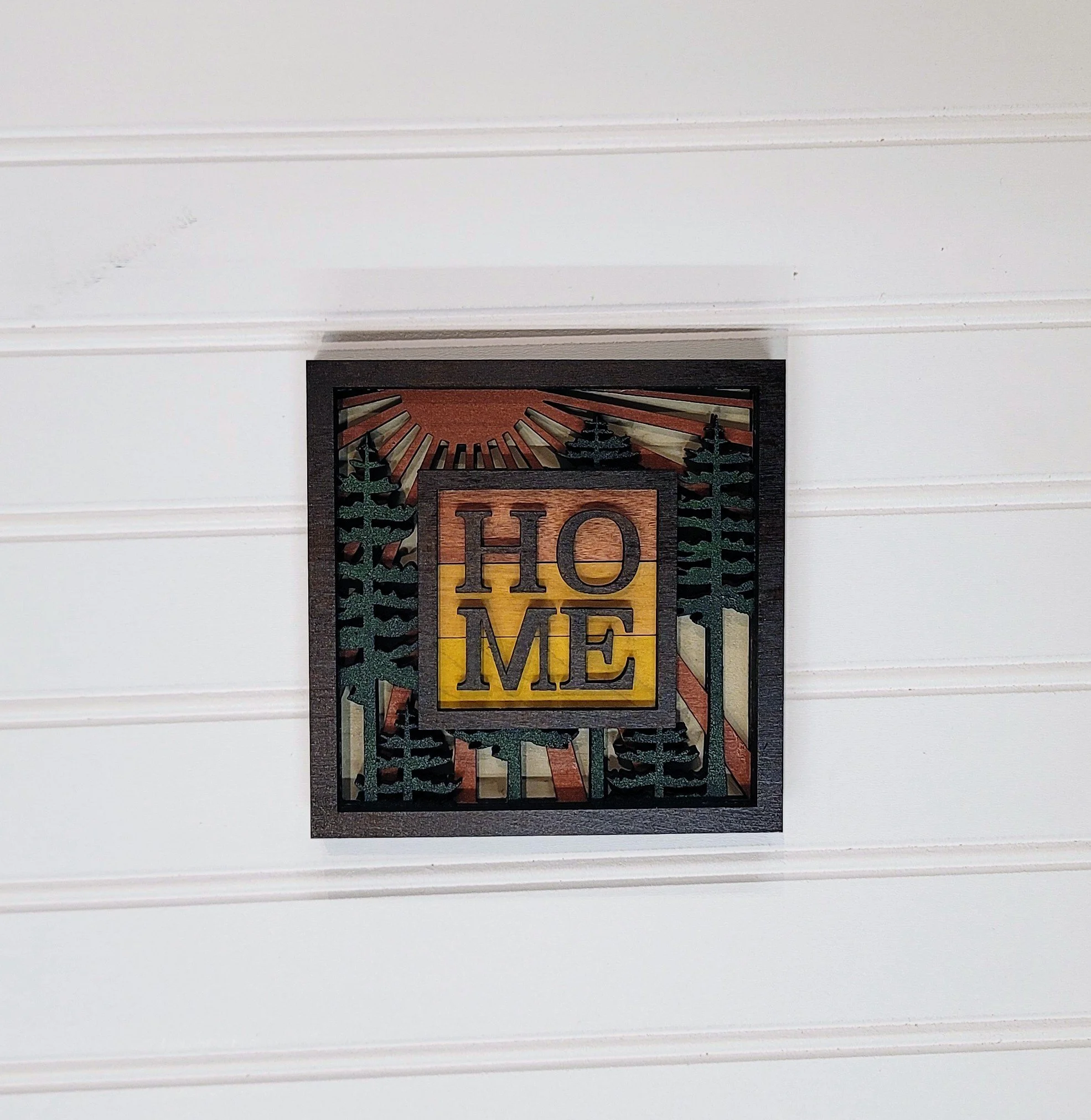 Decorative wall art with the word 'HOME' in a wooden frame, featuring trees, a sun, and a sunburst design.