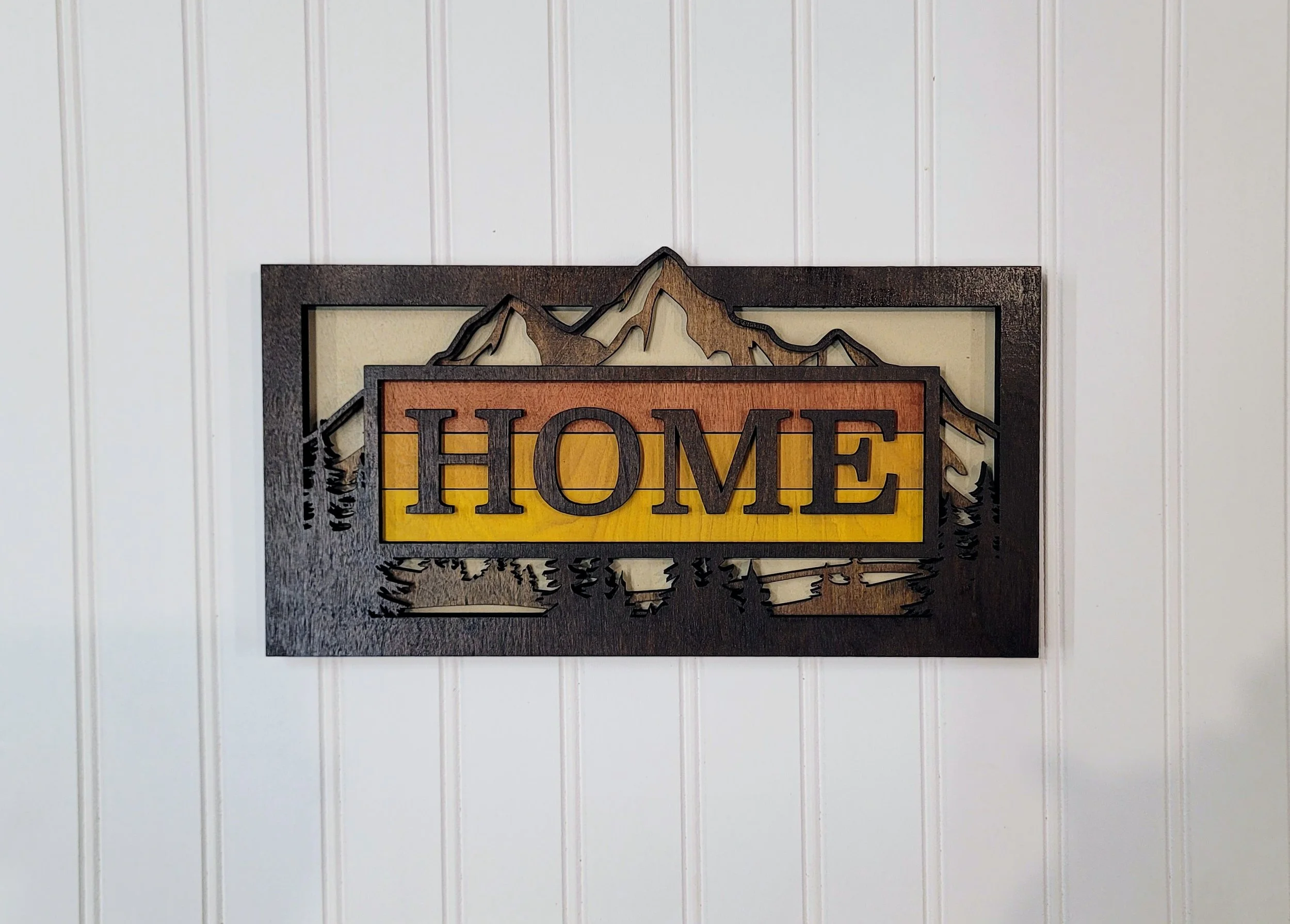 Wooden wall art with mountain and trees design and the word 'HOME' in the center, mounted on a white paneled wall.