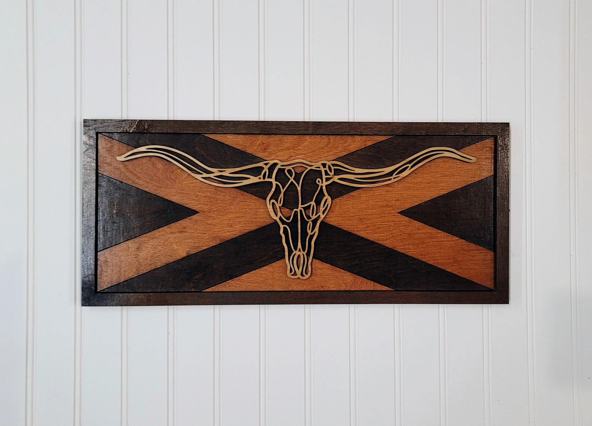 A rectangular wooden wall art piece featuring a minimalist longhorn skull outline overlaying a background of a stylized Confederate flag with a black border.