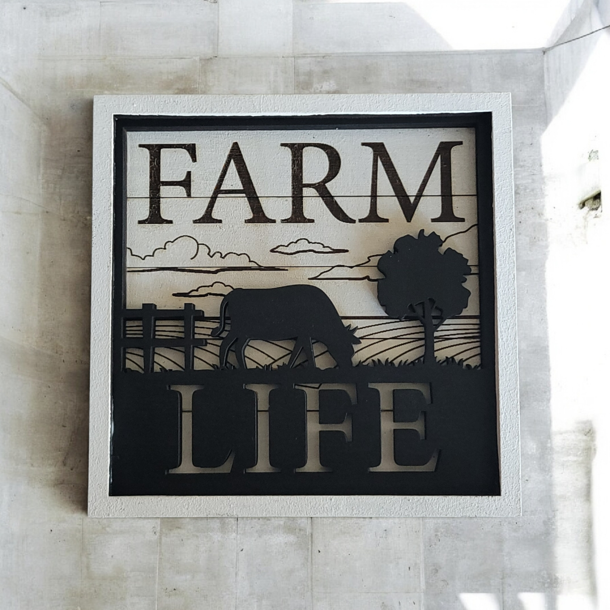 Decorative wall art with the words 'FARM LIFE' and silhouettes of a cow, a tree, clouds, and farmland.