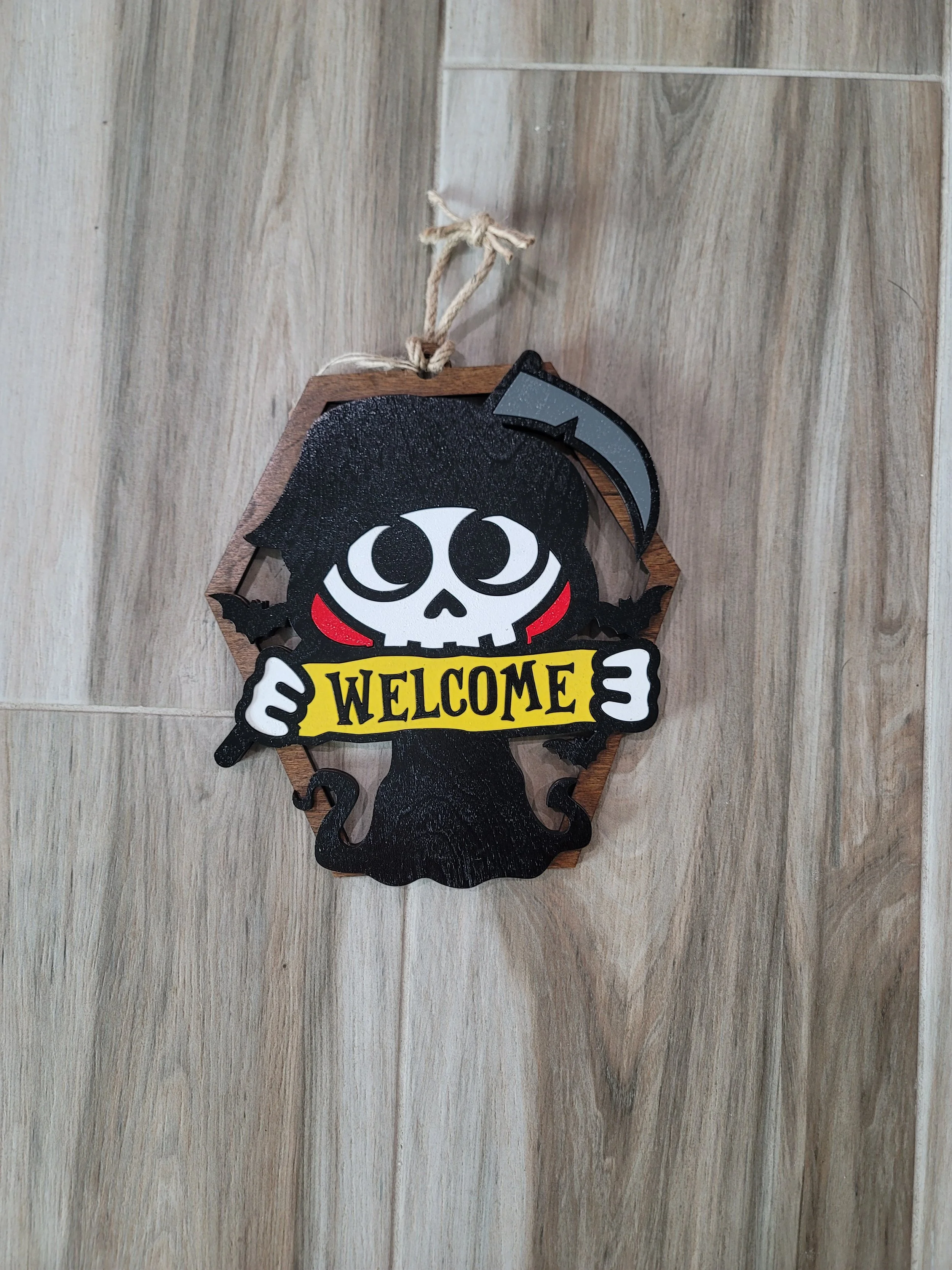 Wooden Halloween-themed wall hanging featuring a black cartoon scythe, a cartoon skull with large eyes, a second smaller skull, and bats, with the word 'WELCOME' in yellow on a banner.