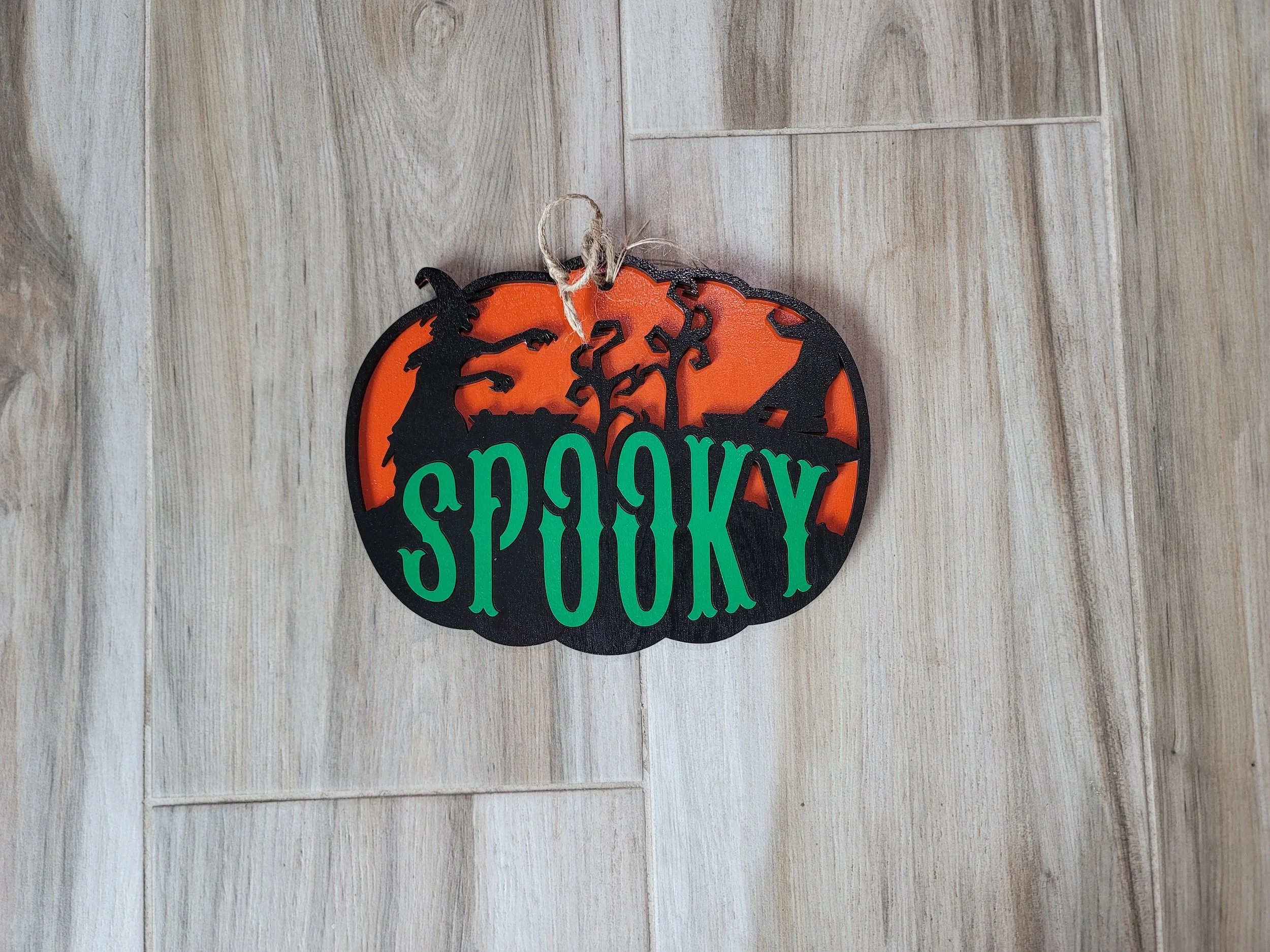 A Halloween-themed sign in the shape of a pumpkin, decorated with a witch hat, a witch silhouette, autumn trees, and the word 'SPOOKY' in green letters, all against an orange background with black outline.