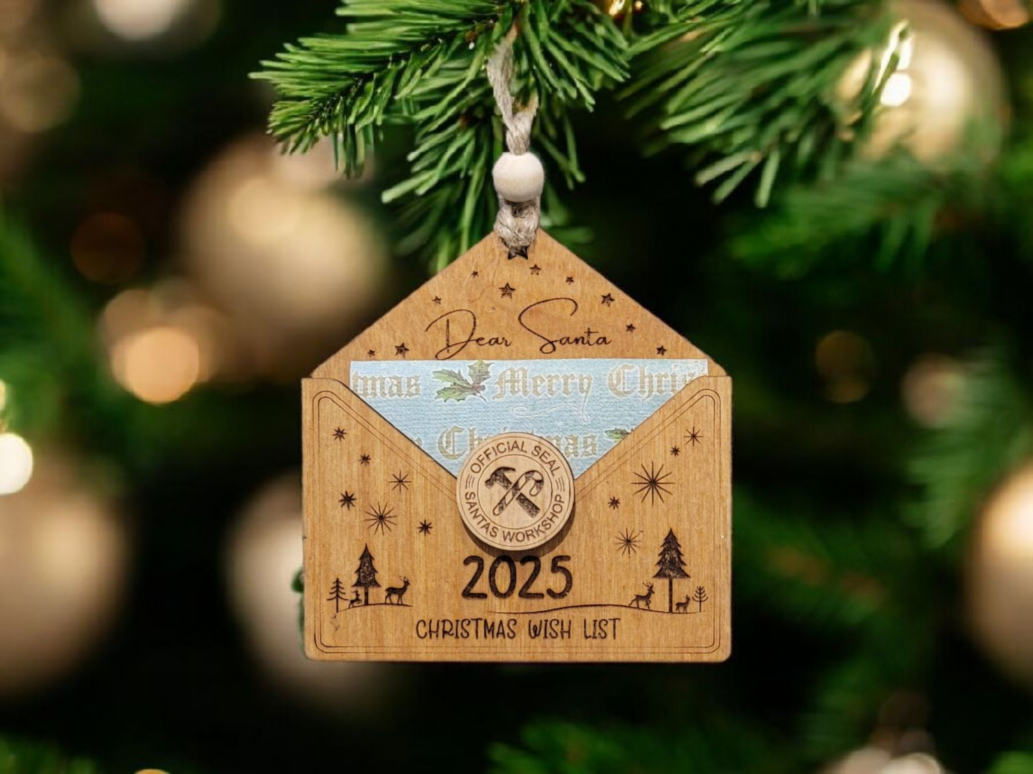 Christmas tree ornament with a wooden house-shaped design, a small blue sticker inside, and a circular seal. The ornament has engraving characters and images including Christmas trees, reindeer, and stars. It reads 'Dear Santa', '2025', and 'Christma