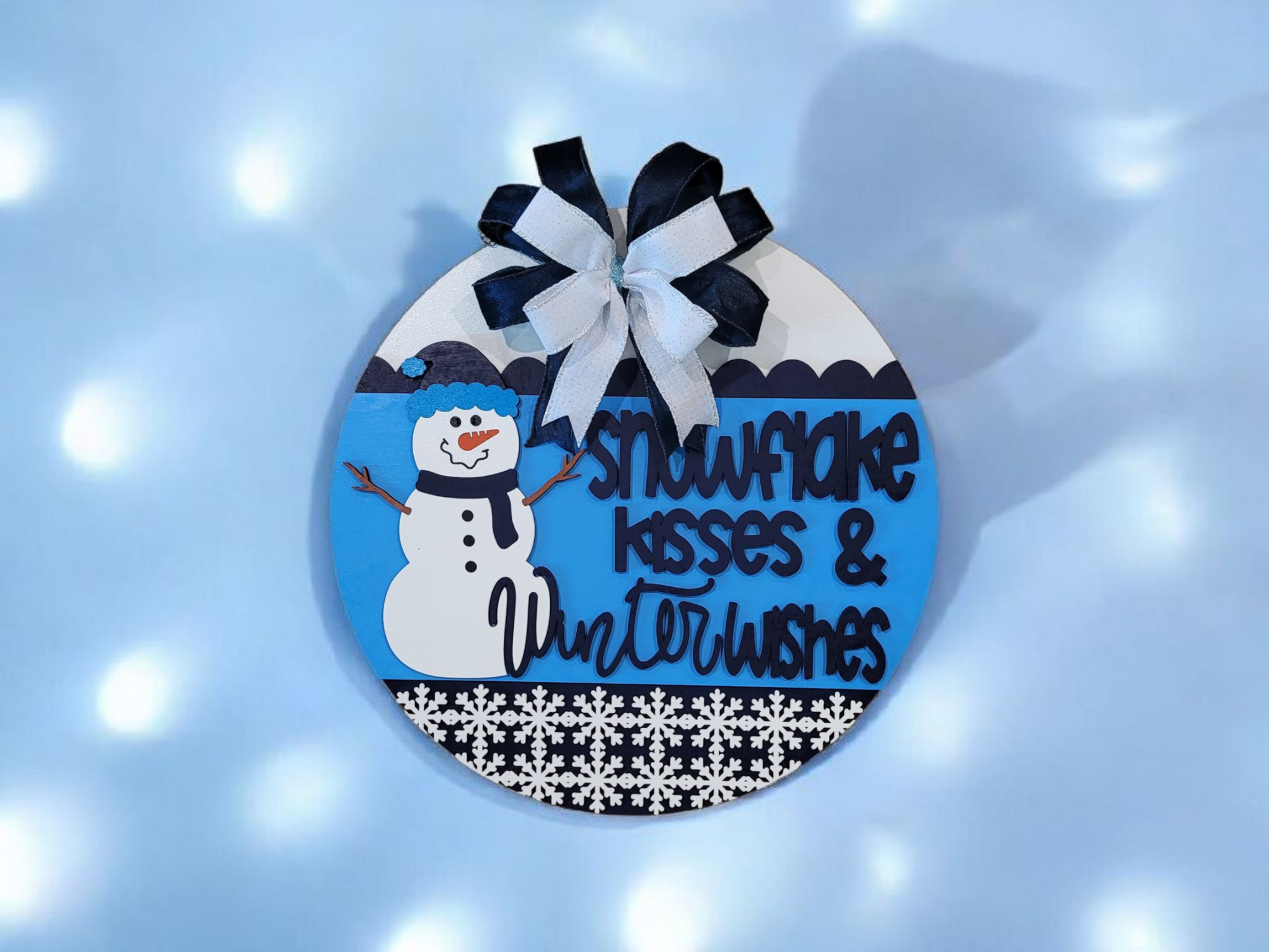 Circular sign with illustrations of a snowman and snowflakes, decorated with a black and white ribbon and clouds, with text that reads "Snowflake kisses & Winter wishes" against a snowy background.