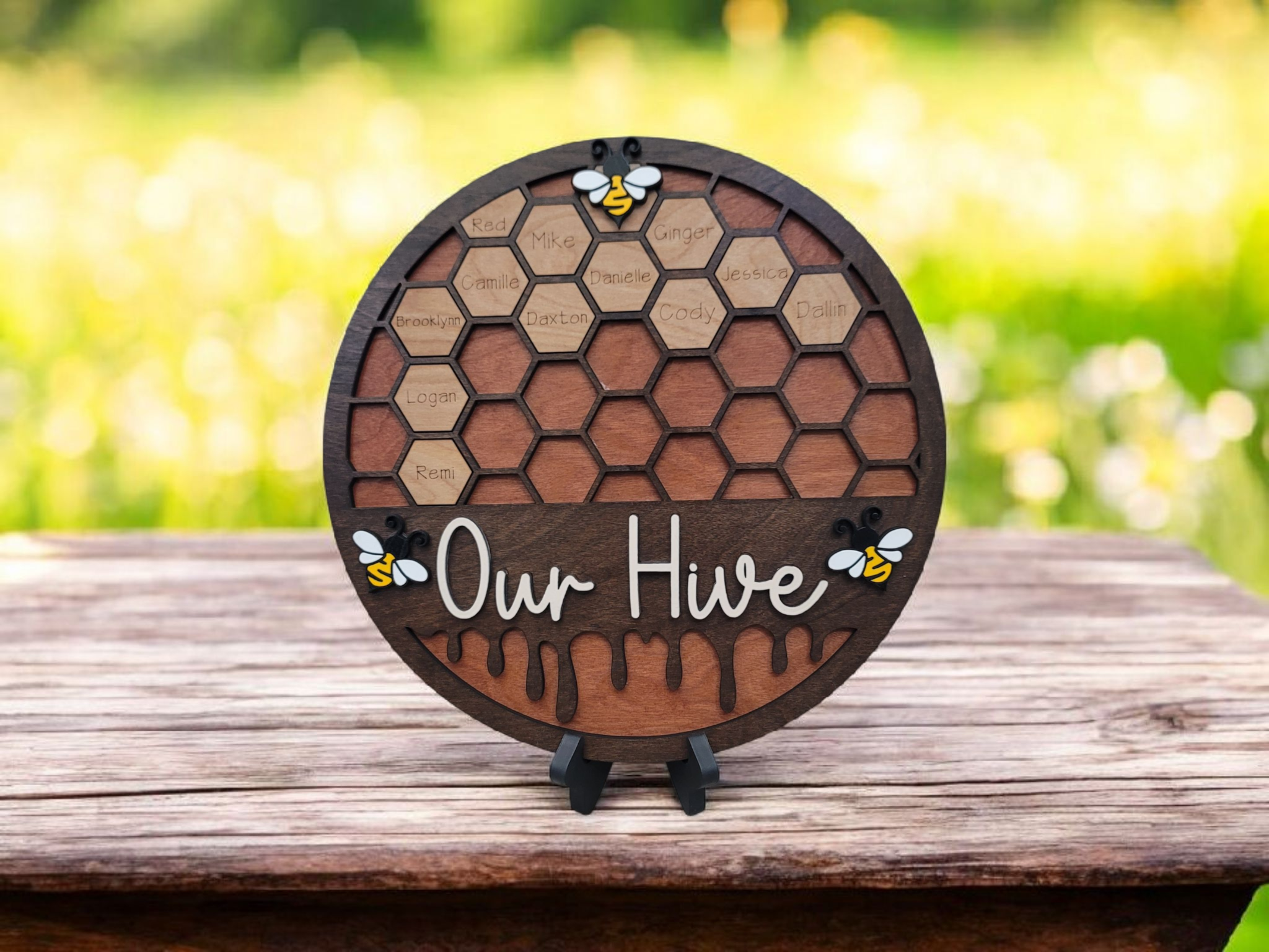 Wooden honey bee house with a honeycomb pattern labeled with names, decorated with bee and honey designs, and the words "Our Hive" written on it, on a wooden surface with a blurred green outdoor background.