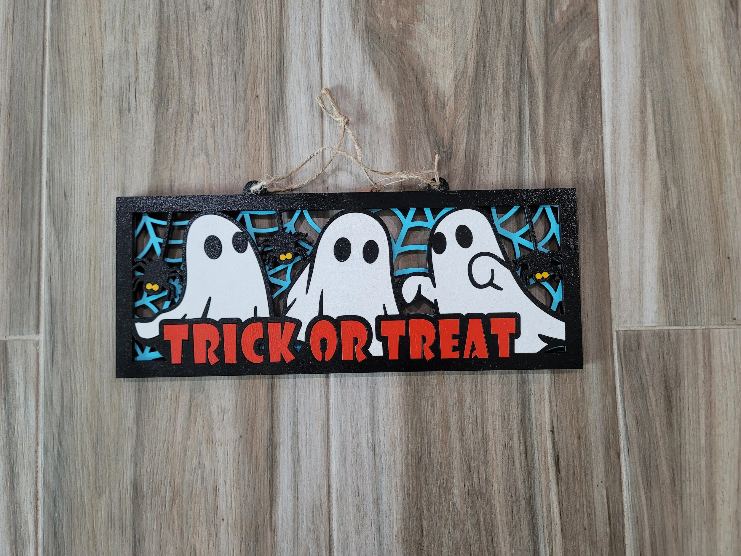 Halloween decoration plaque with ghost figures, black spiders, and blue web background that reads 'Trick or Treat' in red letters.