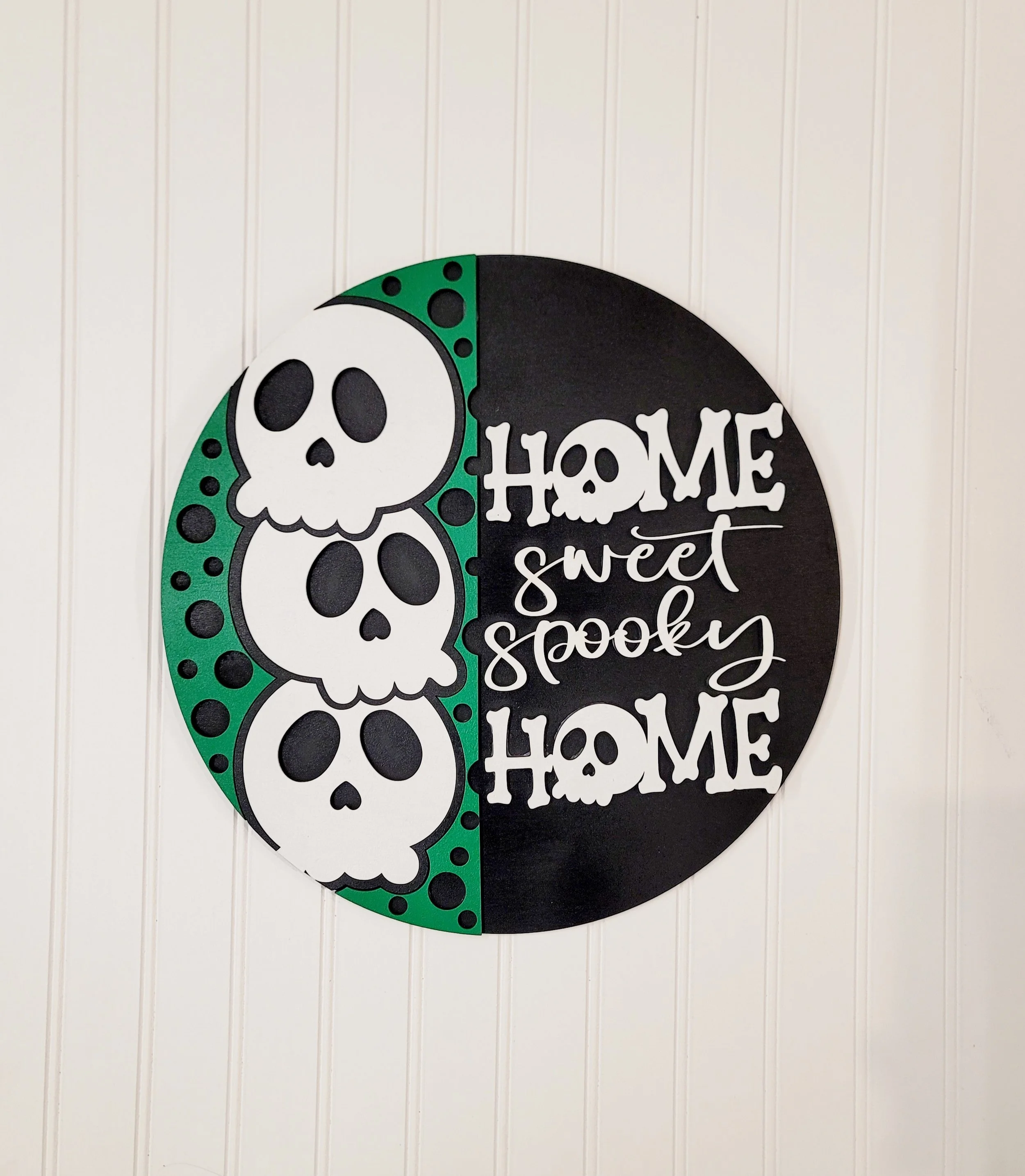 Decorative round sign with three white skulls on the left side and the phrase "Home sweet spooky home" on the right side in white font on a black background, mounted on a white paneled wall.