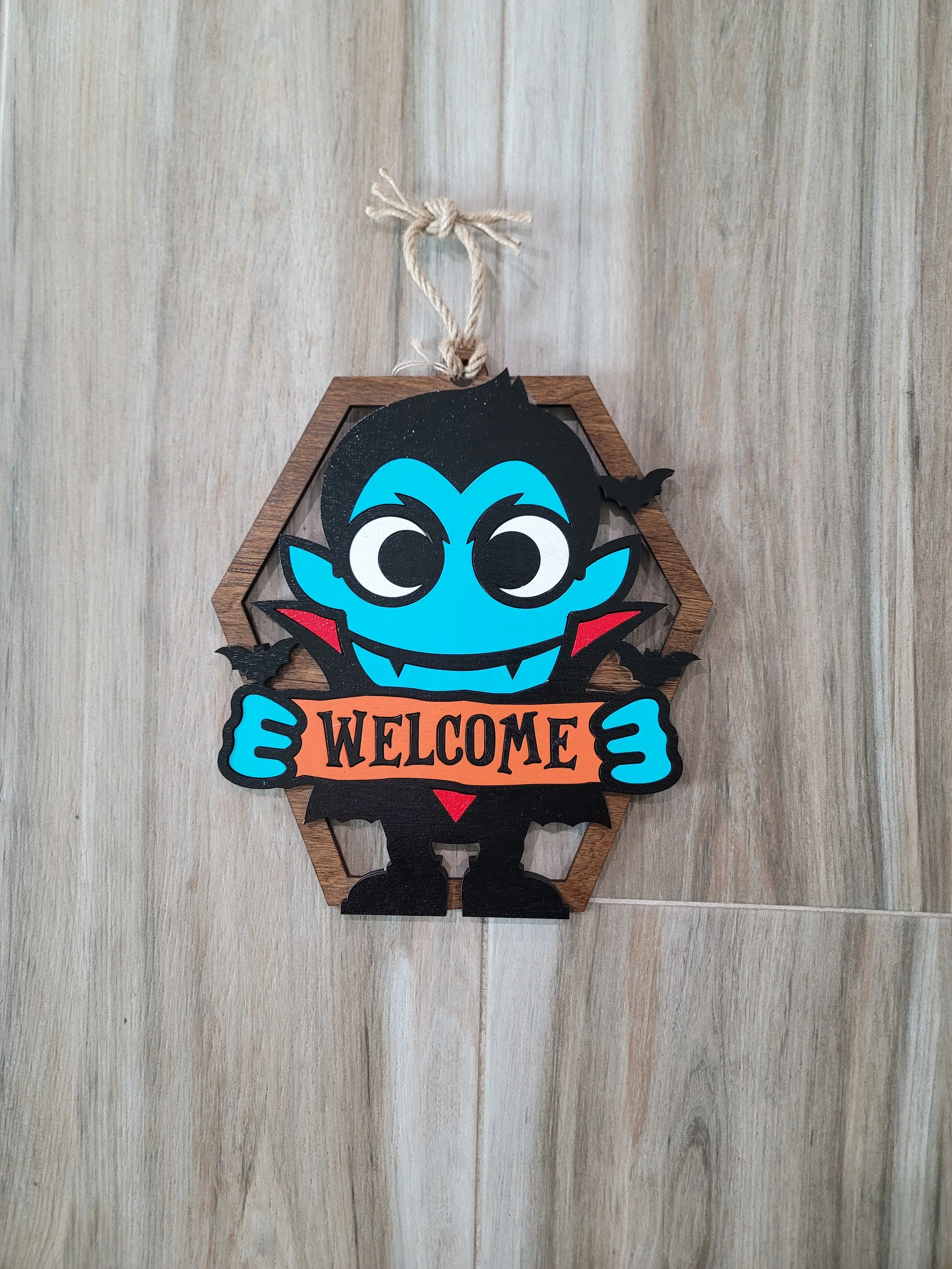 A Halloween-themed welcome sign featuring a cartoon vampire holding a sign with the word 'WELCOME'. The vampire has blue skin, black hair, fangs, red lips, and is surrounded by small black bats. The sign is mounted on a wooden hexagonal frame with a 