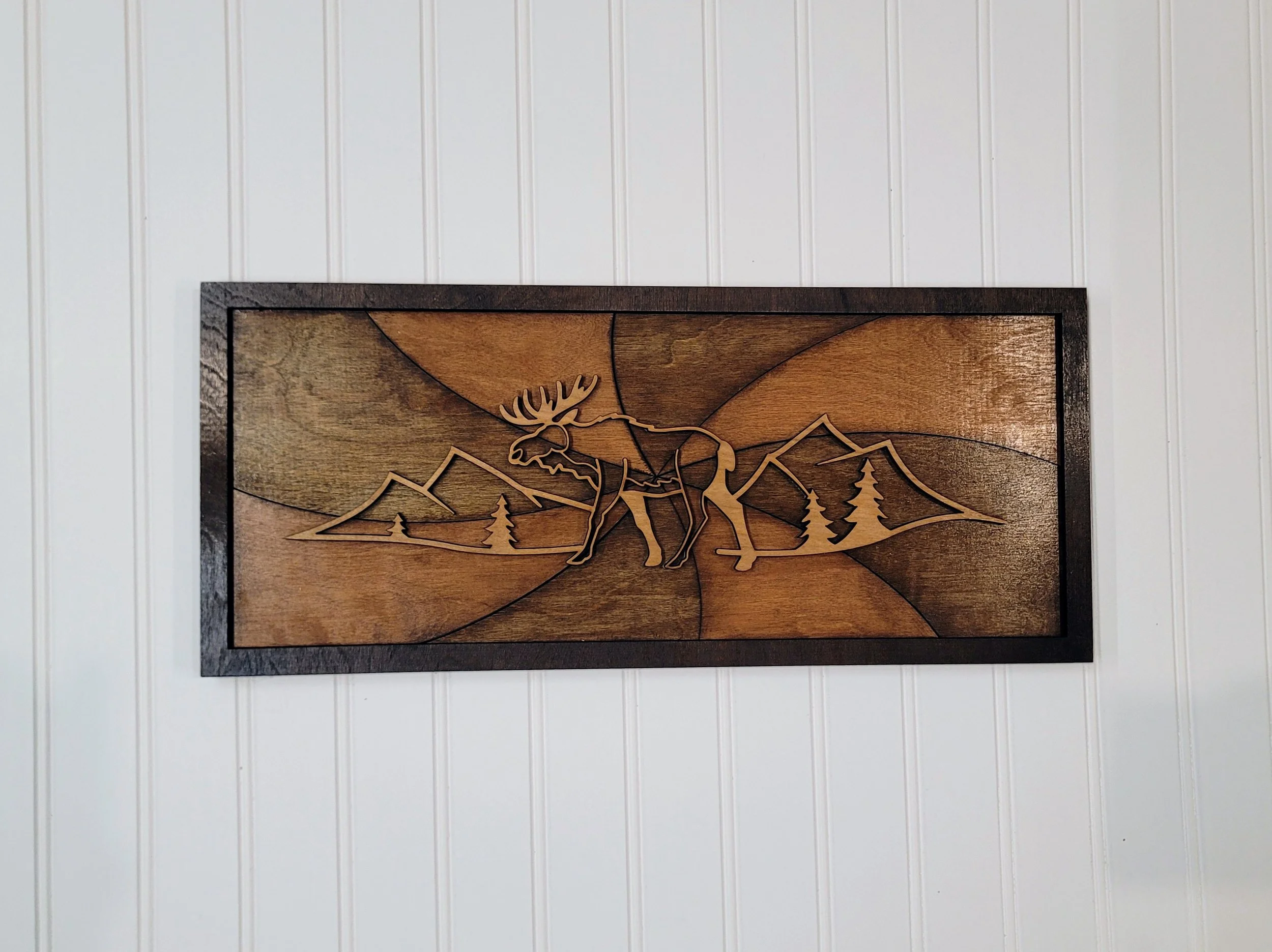 Wooden wall art featuring a moose with mountain and pine tree landscape background.