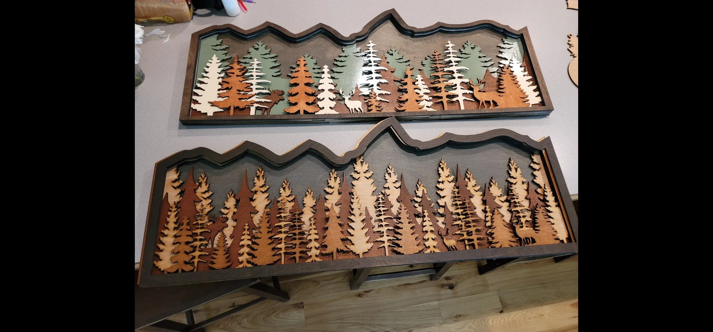 Two wooden framed shadow boxes depicting layered forest scenes with trees and deer, set on a light-colored table.