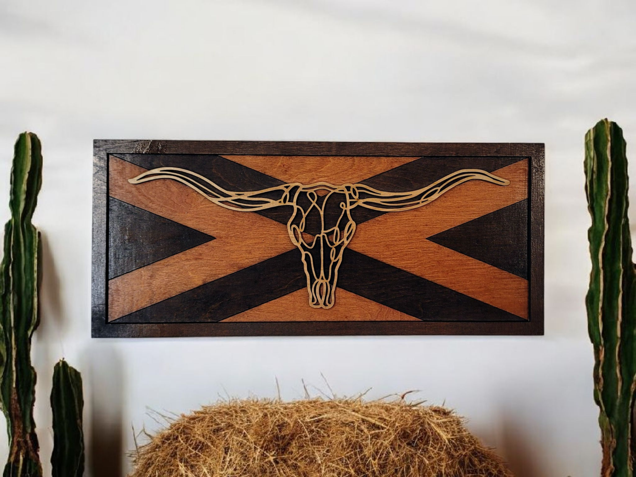 Wooden wall art featuring a longhorn skull design with a southwestern geometric background, flanked by two tall cacti on white walls, with hay in front.