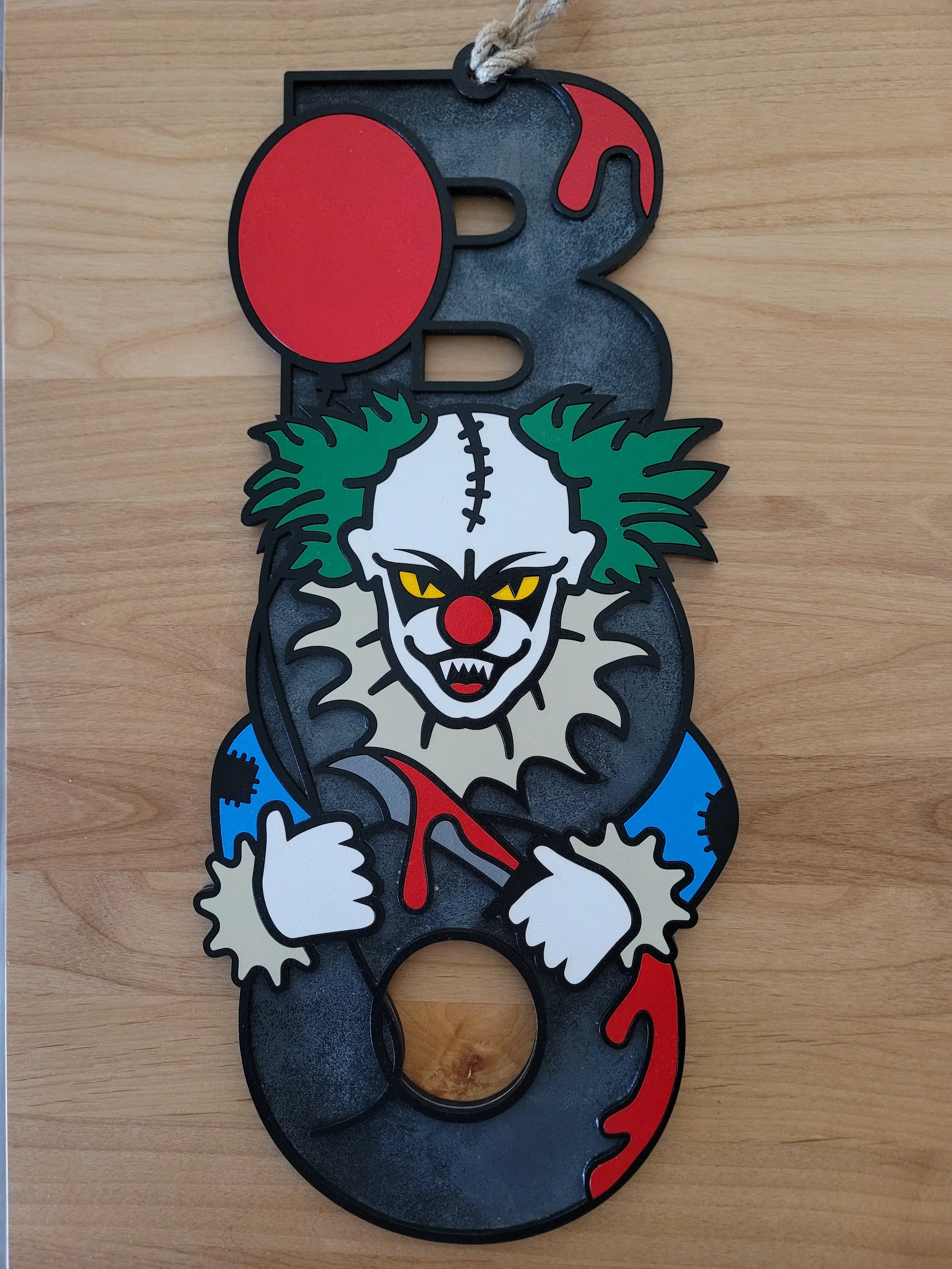 A decorative item shaped like the letter 'B' featuring a sinister clown with yellow eyes, green hair, red nose, white face, and sharp teeth, holding a blood-stained weapon.