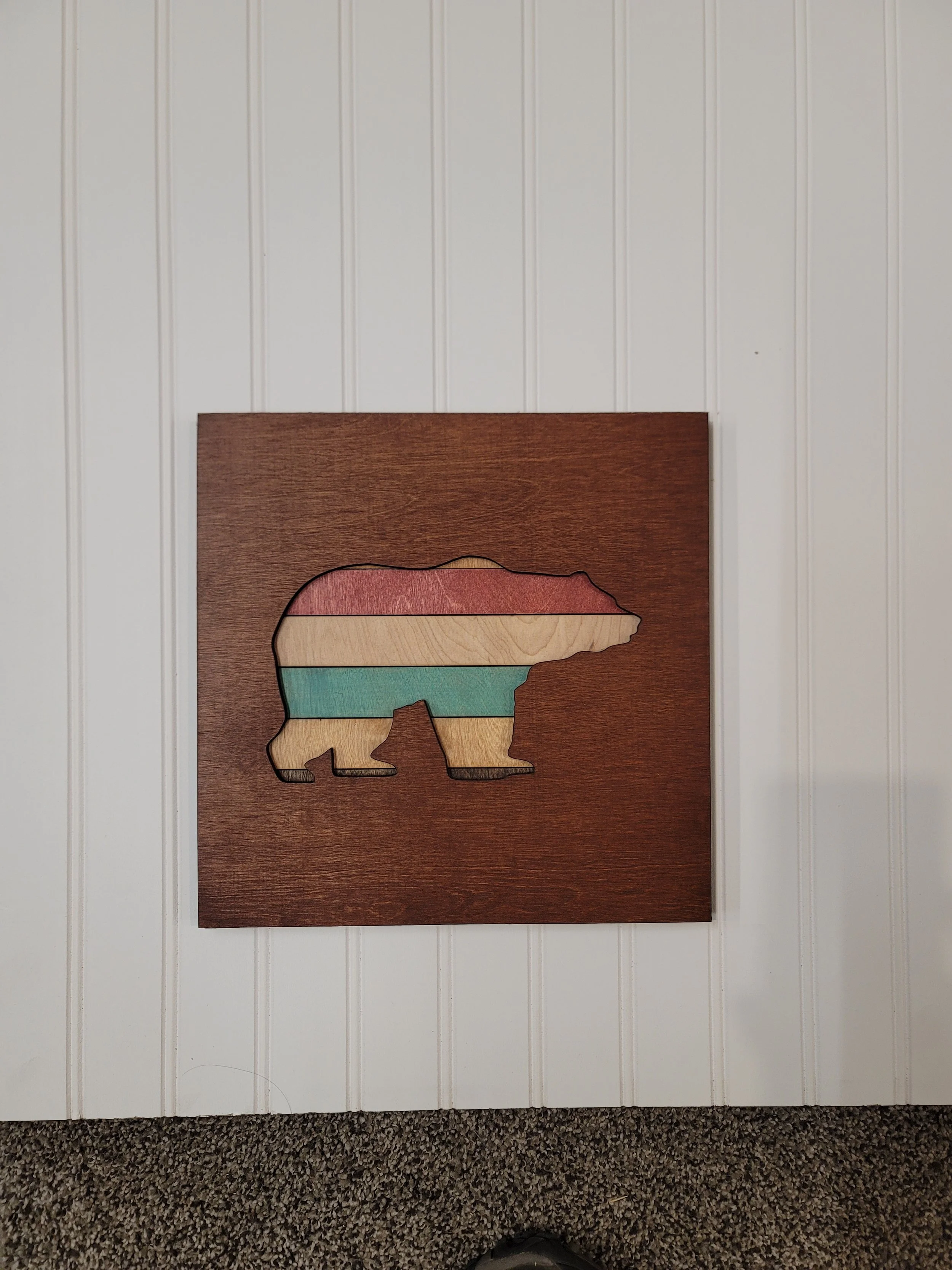 Wooden wall art featuring a bear silhouette filled with striped pastel colors, mounted on white paneled wall.