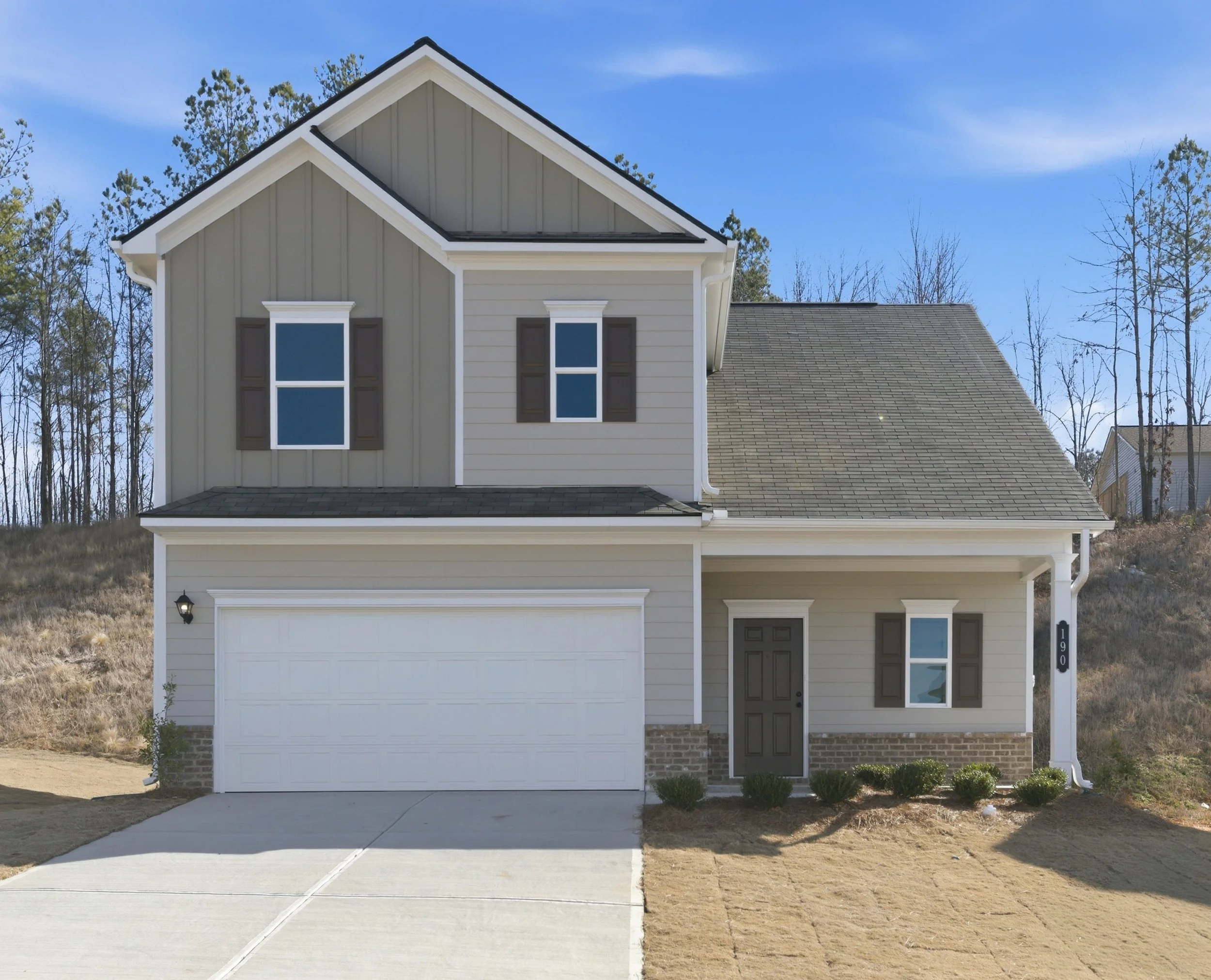 Two-story house with gray and beige siding, black shutters, white trim, and a white garage door. The house has a small front yard with some bushes, a concrete driveway, and a black front door. The house number 190 is on the side post. In the backgrou